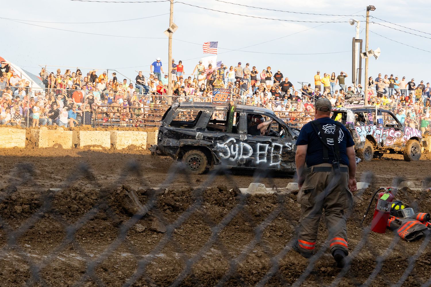 Wide shot of a demolition derby with damaged vehicles competing in muddy conditions.