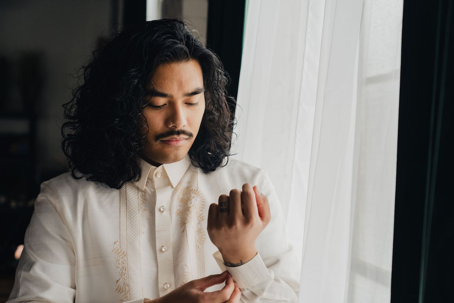 A person with dark curly hair wearing a white shirt looks down thoughtfully while sitting near a window.