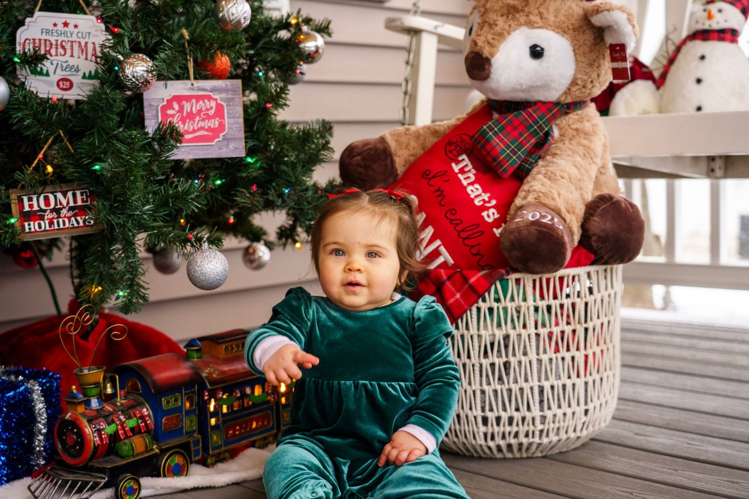 Holiday scene with a baby in green velvet next to a Christmas tree and decorative teddy bear.