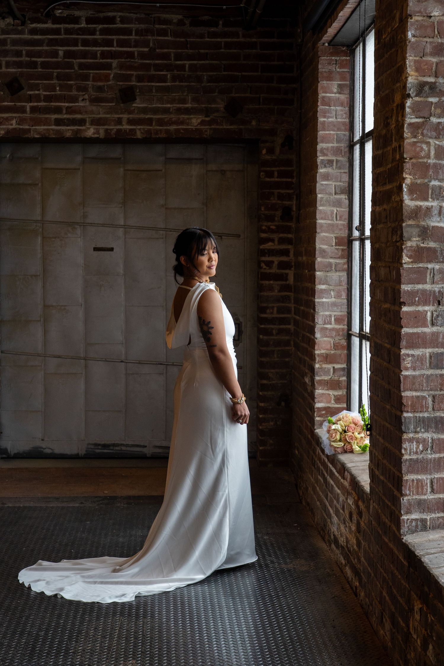 A bride in a flowing white dress poses in a rustic brick industrial space near large windows.