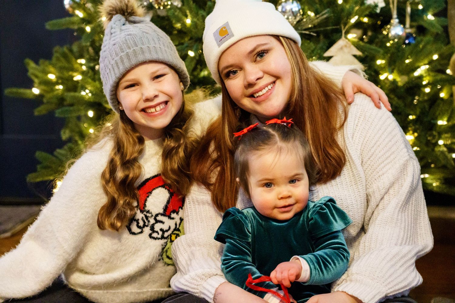 Family members share warm holiday moments in front of a Christmas tree with twinkling lights in the background.