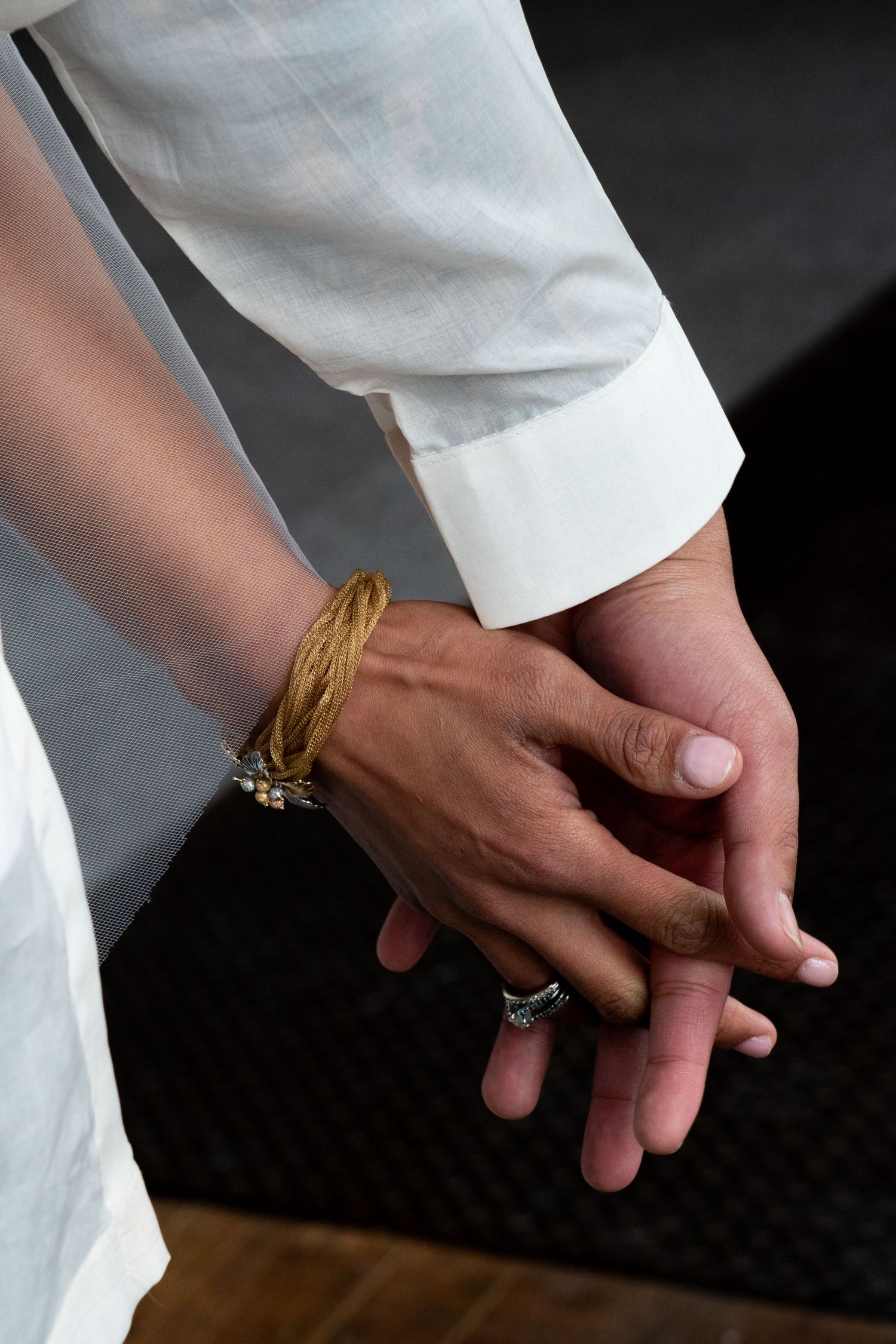 Close-up of hands wearing white sleeves, gold bracelet and rings against dark background.
