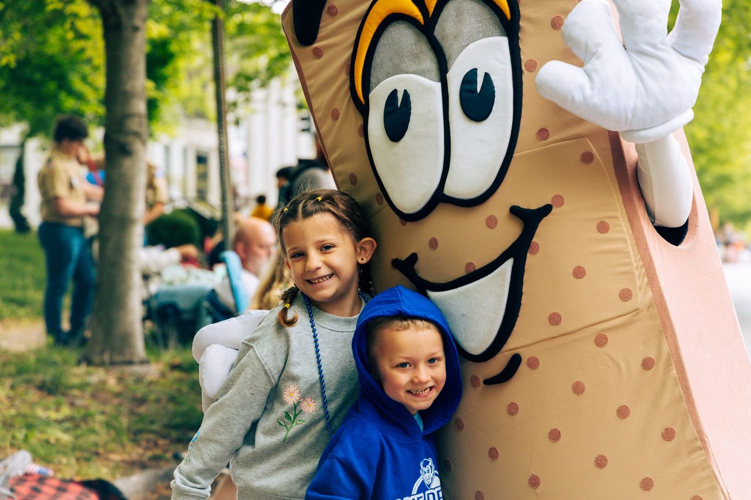Children posing with large mascot costume character at outdoor community event.