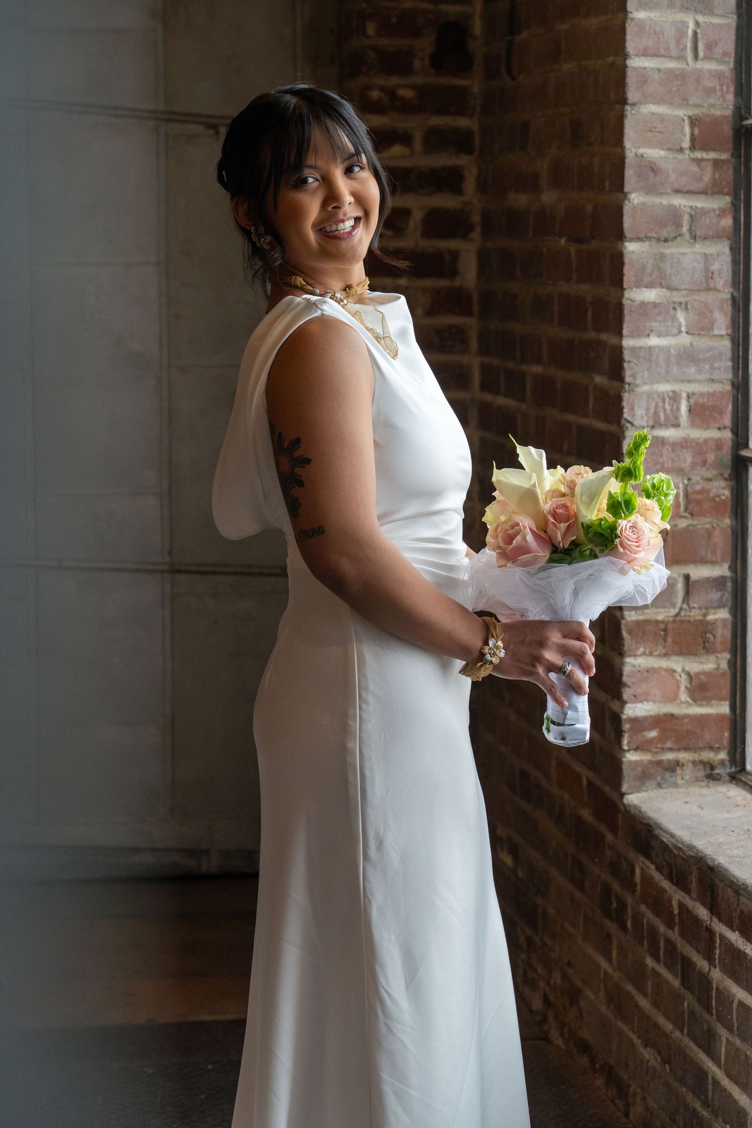 A person in a white wedding dress smiles while standing against a rustic brick wall with natural lighting.