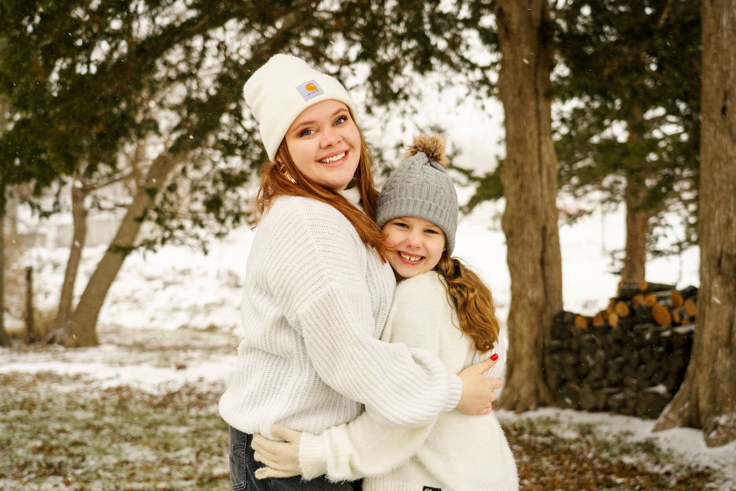 A tender winter moment captured with warm embraces and matching white knit hats against snowy trees.
