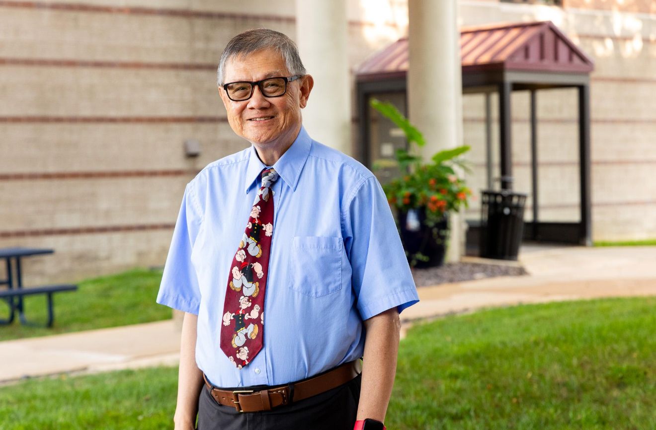 Professional portrait in a light blue dress shirt and burgundy tie against a stone building backdrop.