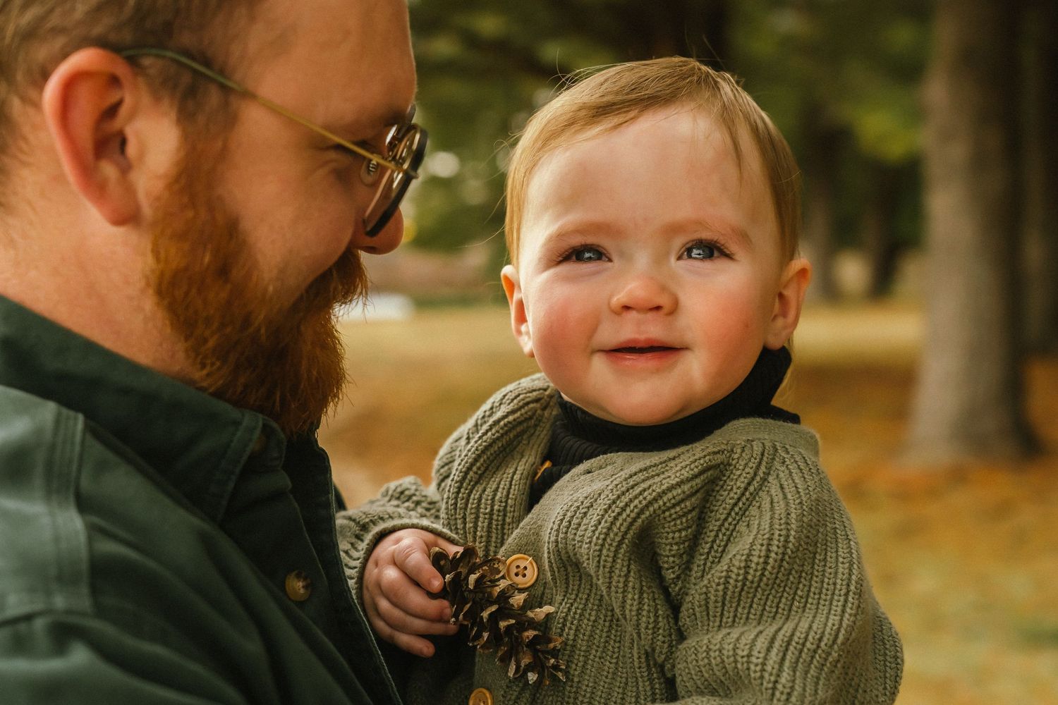 A toddler in a green knit sweater smiles while being held outdoors during autumn.