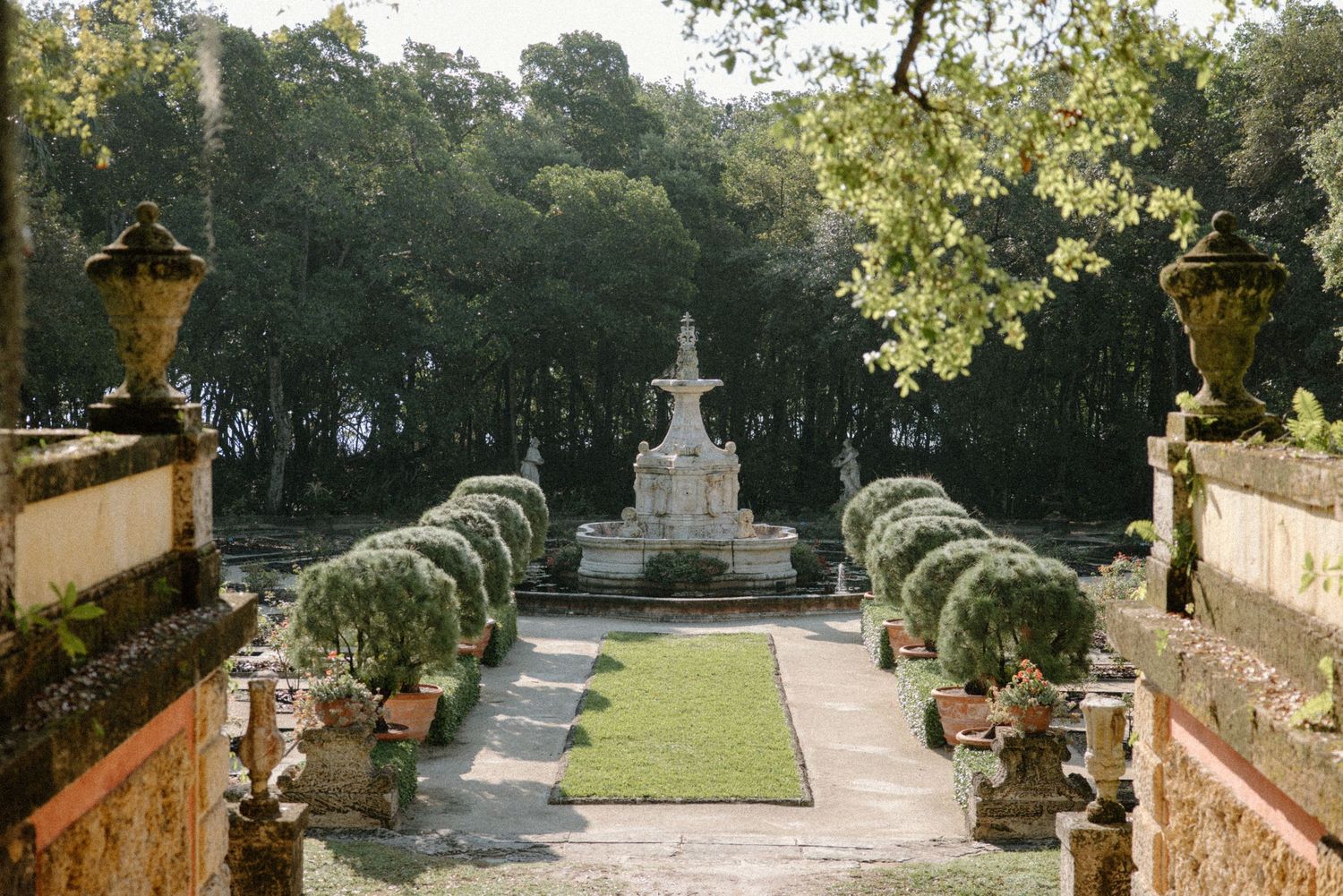 Elegant Italian garden with stone fountain, manicured hedges, and classical urns framed by tall trees.