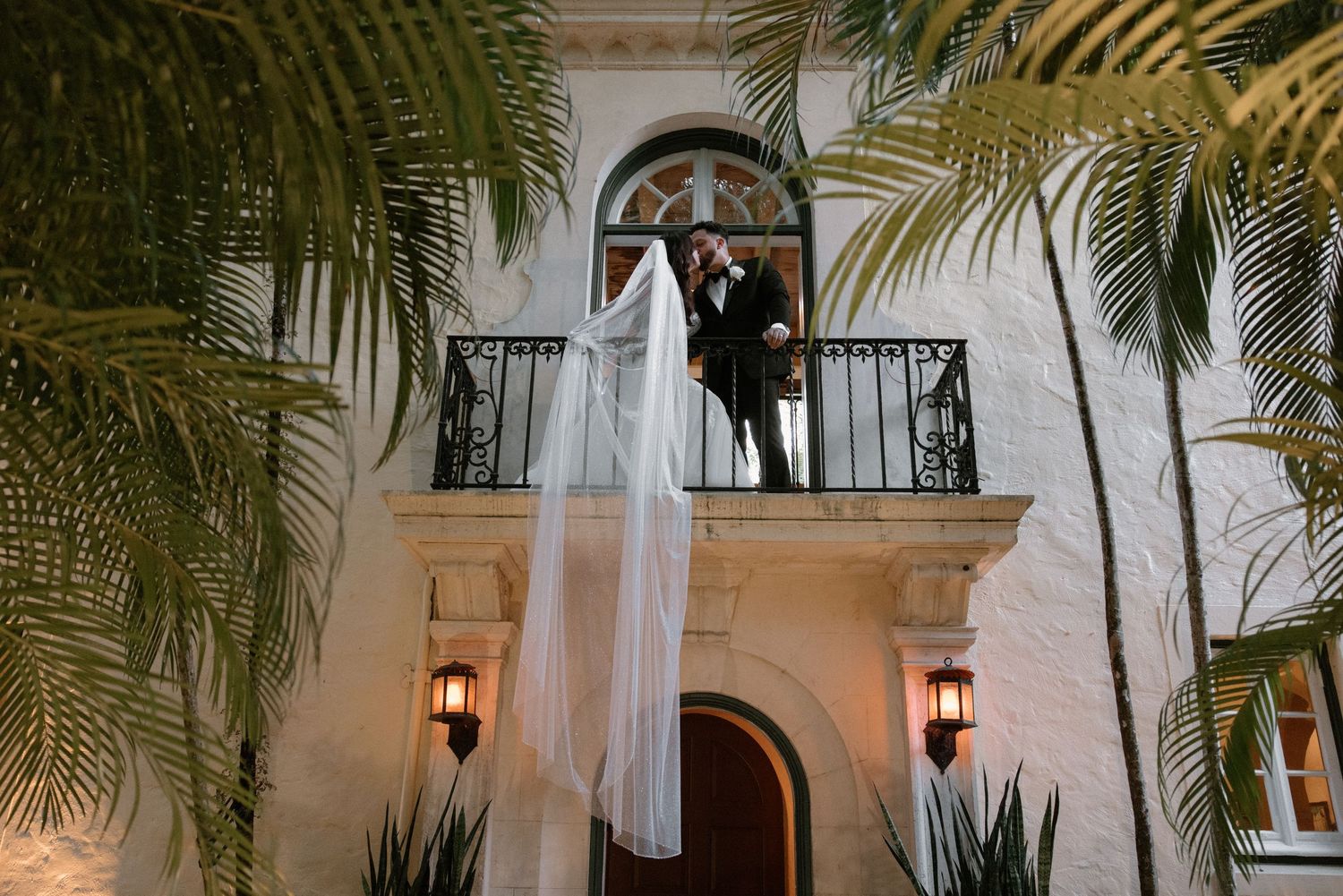 Wedding dress hangs elegantly from balcony of Mediterranean-style villa with palm trees and glowing lanterns at dusk.