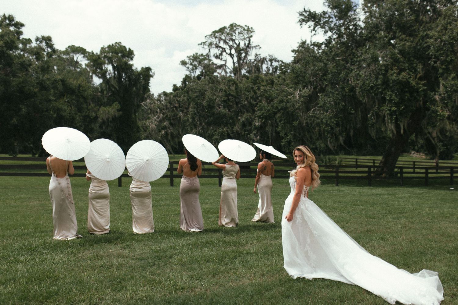 A row of people in pink dresses holding white parasols stand in a grassy field on a sunny day.