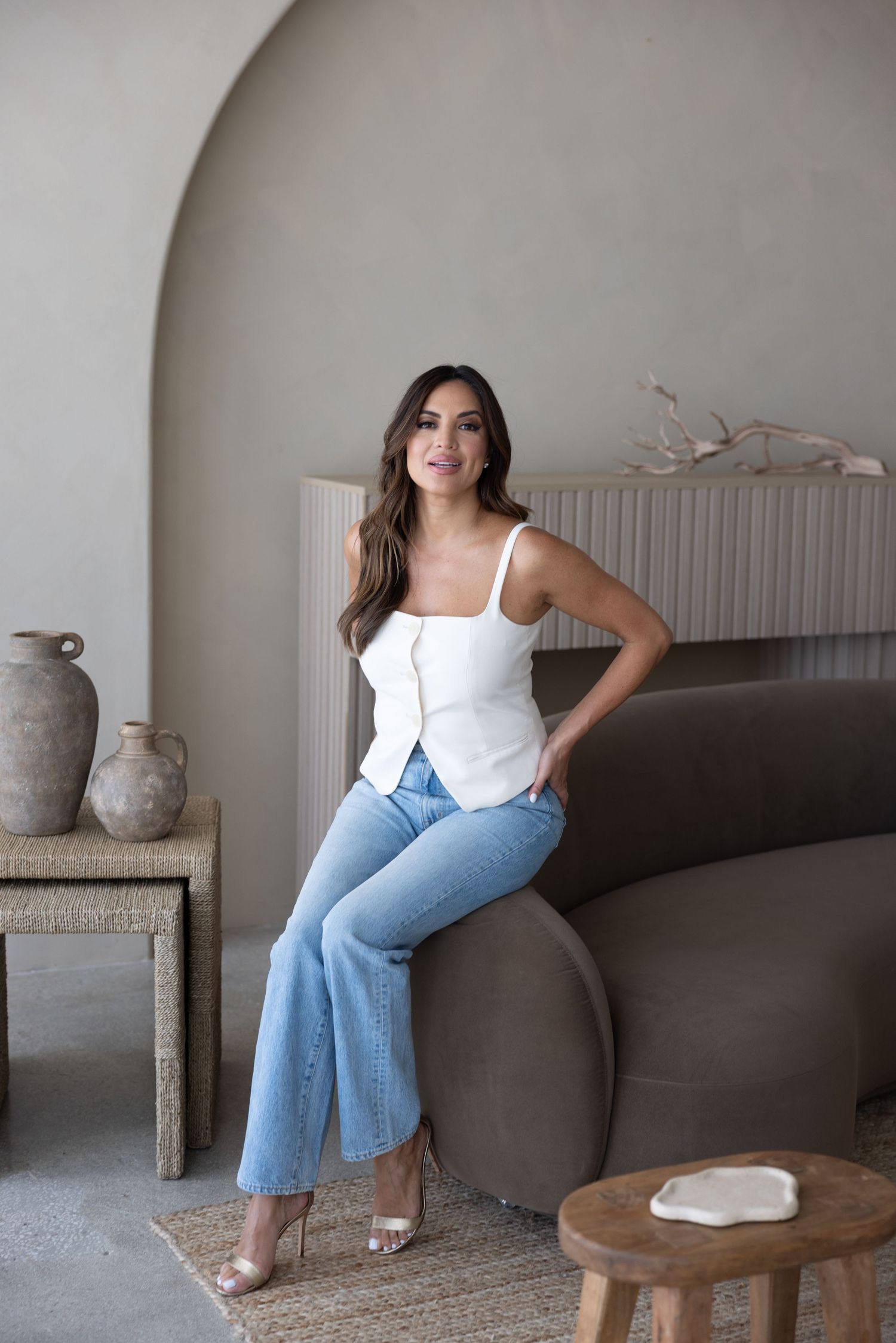 Woman in white tank top and blue wide-leg jeans sitting on gray curved sofa in minimalist neutral-toned living room.