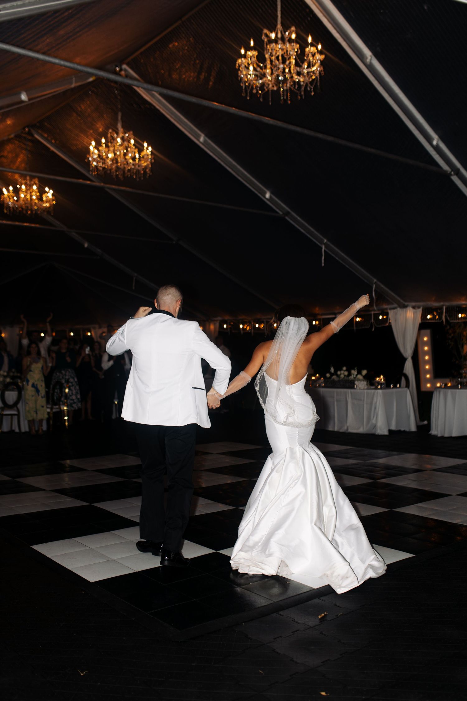 A couple in formal wedding attire dances on a black and white checkered dance floor under elegant chandeliers.