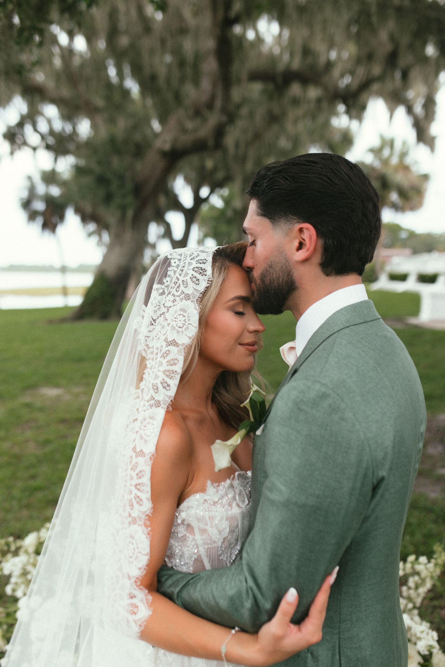 A tender wedding moment captured outdoors under a tree with the bride in lace and groom in sage green suit.