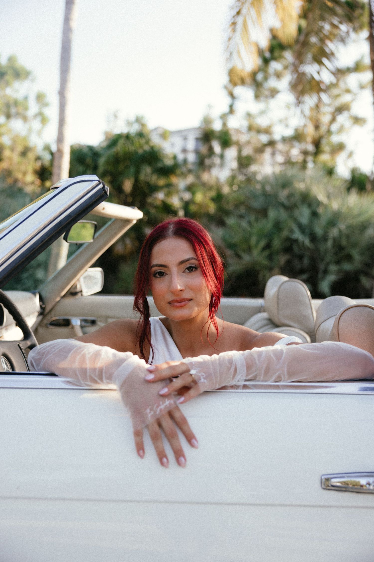 A person with red hair poses while reclining in a white vintage car on a sunny day.