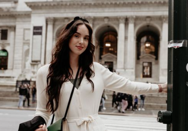 A person in a white dress poses against a classical building exterior with columns.