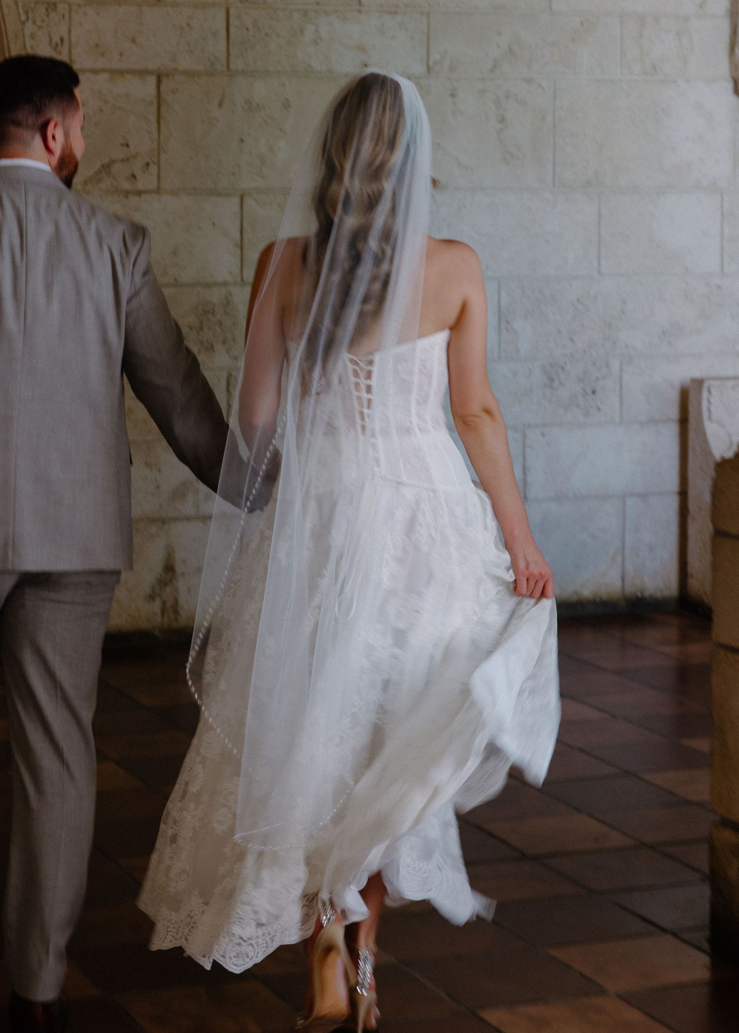 A couple in wedding attire walks together across a stone-tiled floor in a historic building setting.