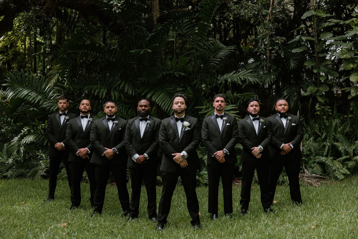 Wedding party in black tuxedos poses outdoors in a garden setting with greenery in the background.