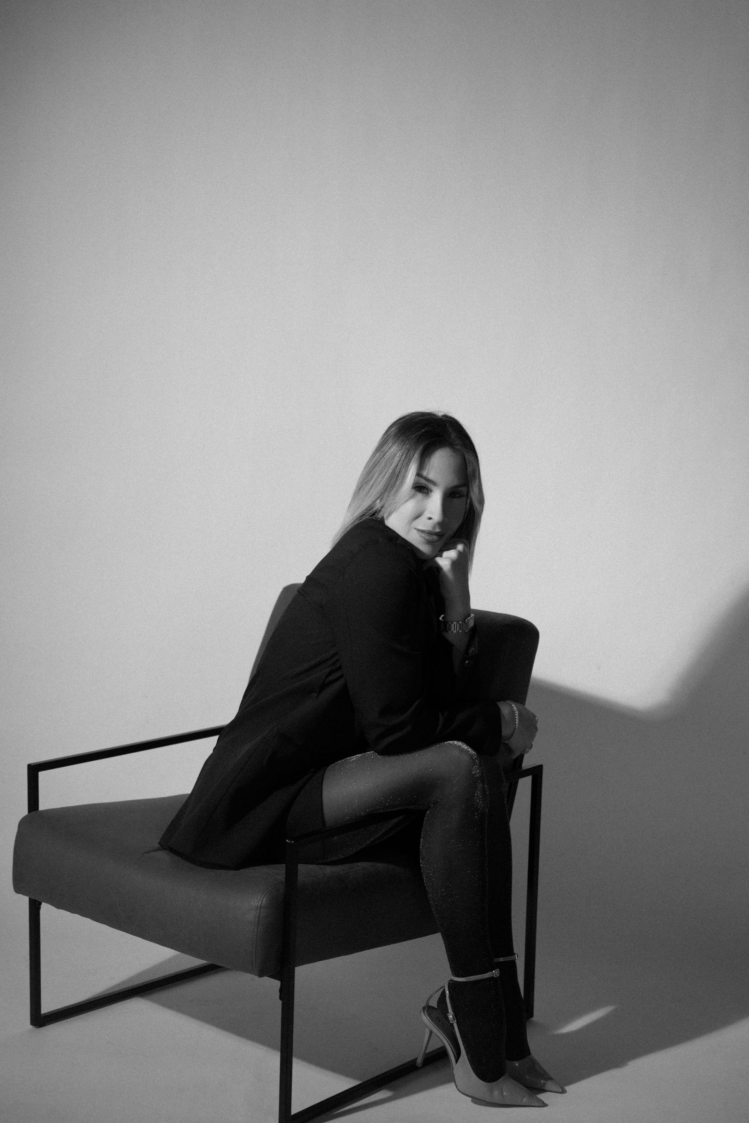 Black and white portrait of a woman in a blazer sitting on a modern chair, looking over her shoulder elegantly.