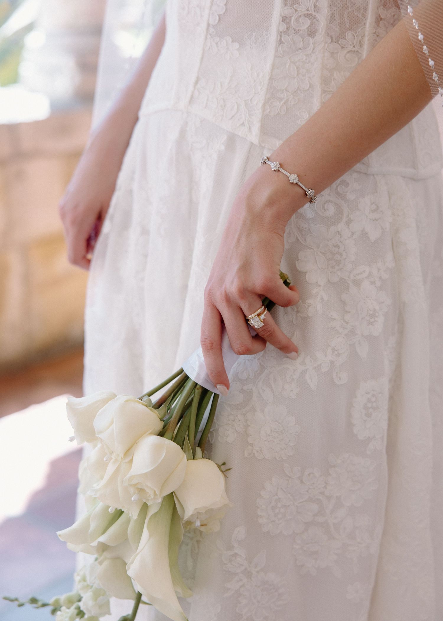 Close-up of bride in white lace dress holding elegant white calla lily bouquet with delicate bracelet and ring details.