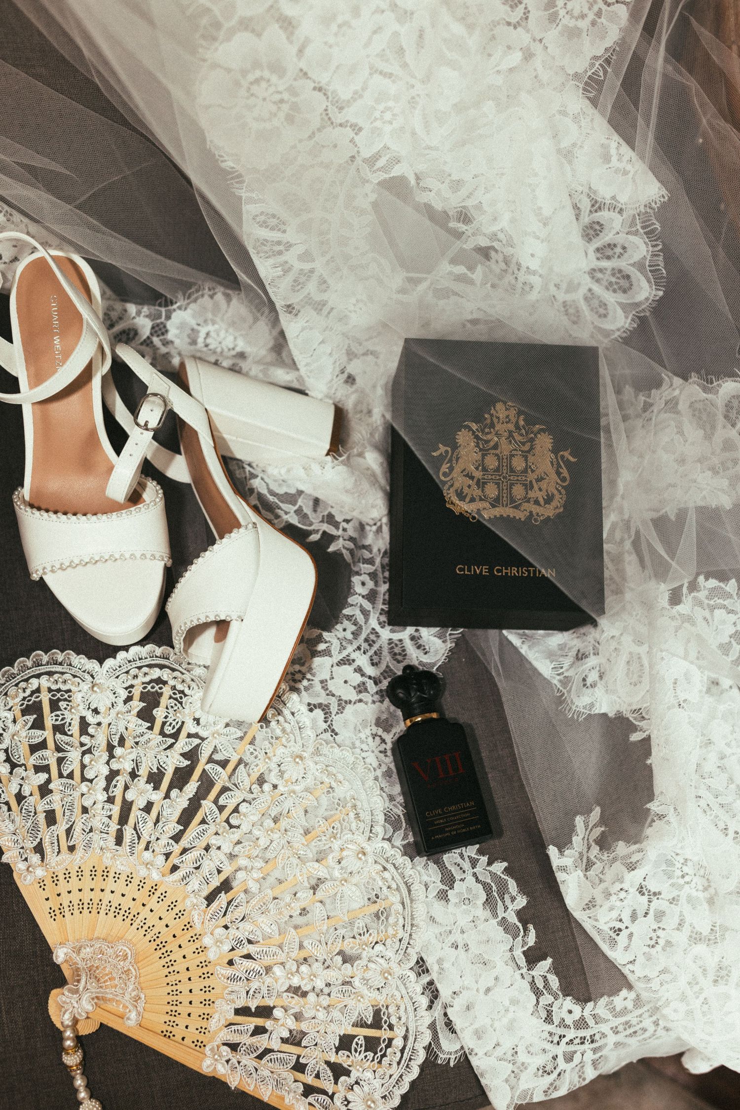 White lace wedding veil displayed with elegant bridal shoes and a decorative gold folding fan against a dark background.