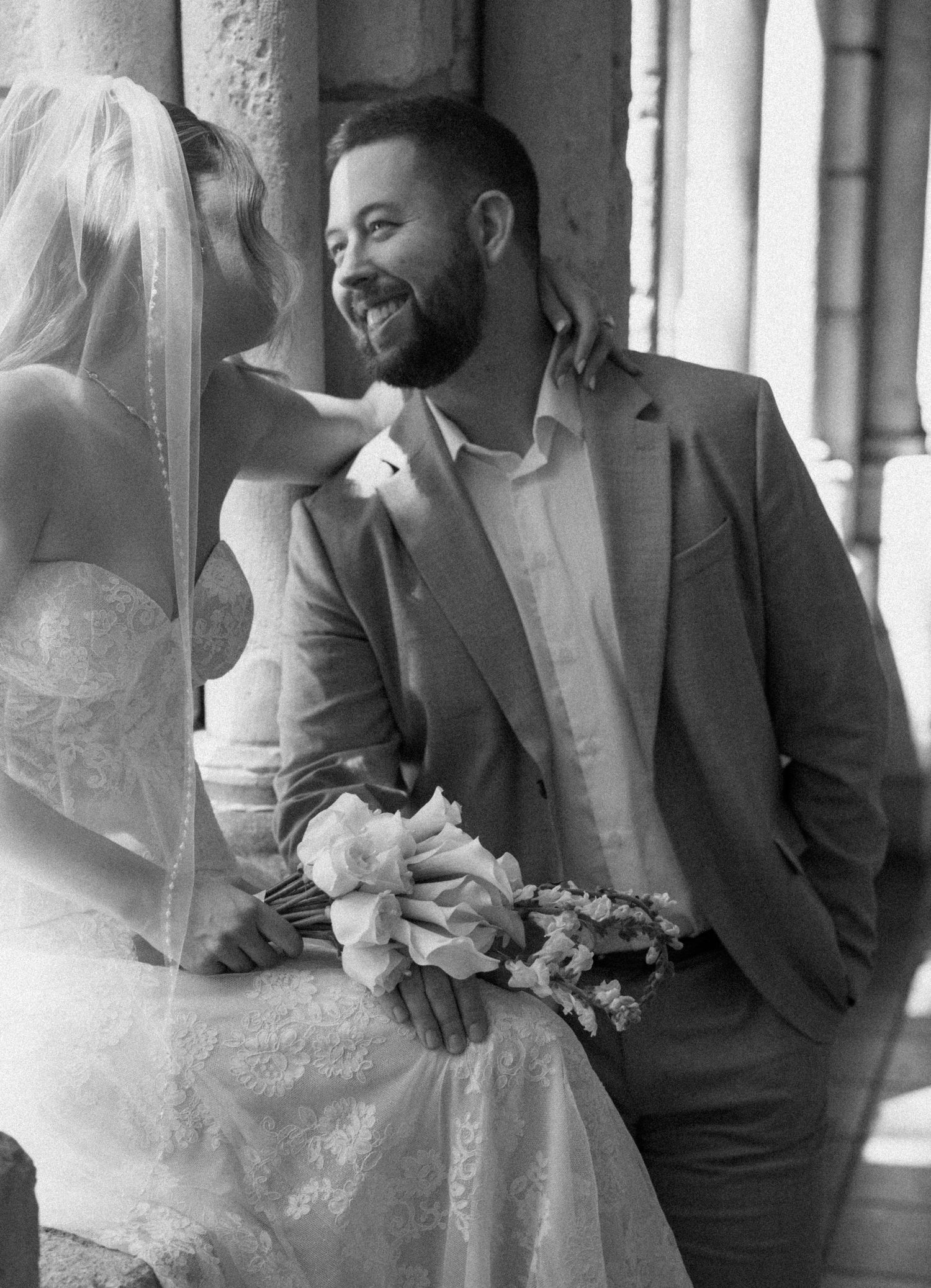 Black and white wedding photo showing bride and groom sharing intimate moment in elegant archway corridor.