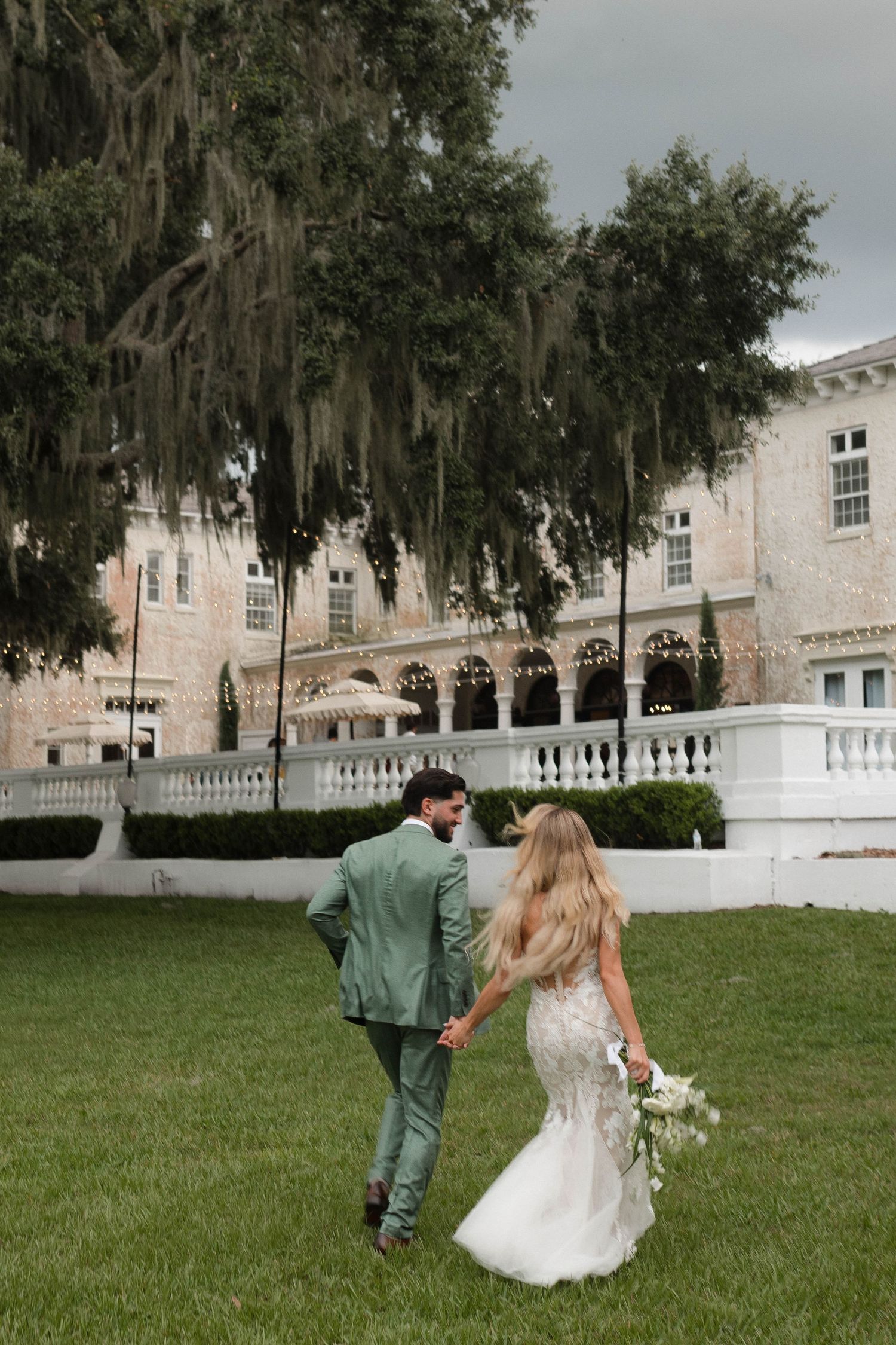 A couple walks across a grassy lawn in front of a historic white building with Spanish moss draped oak trees.