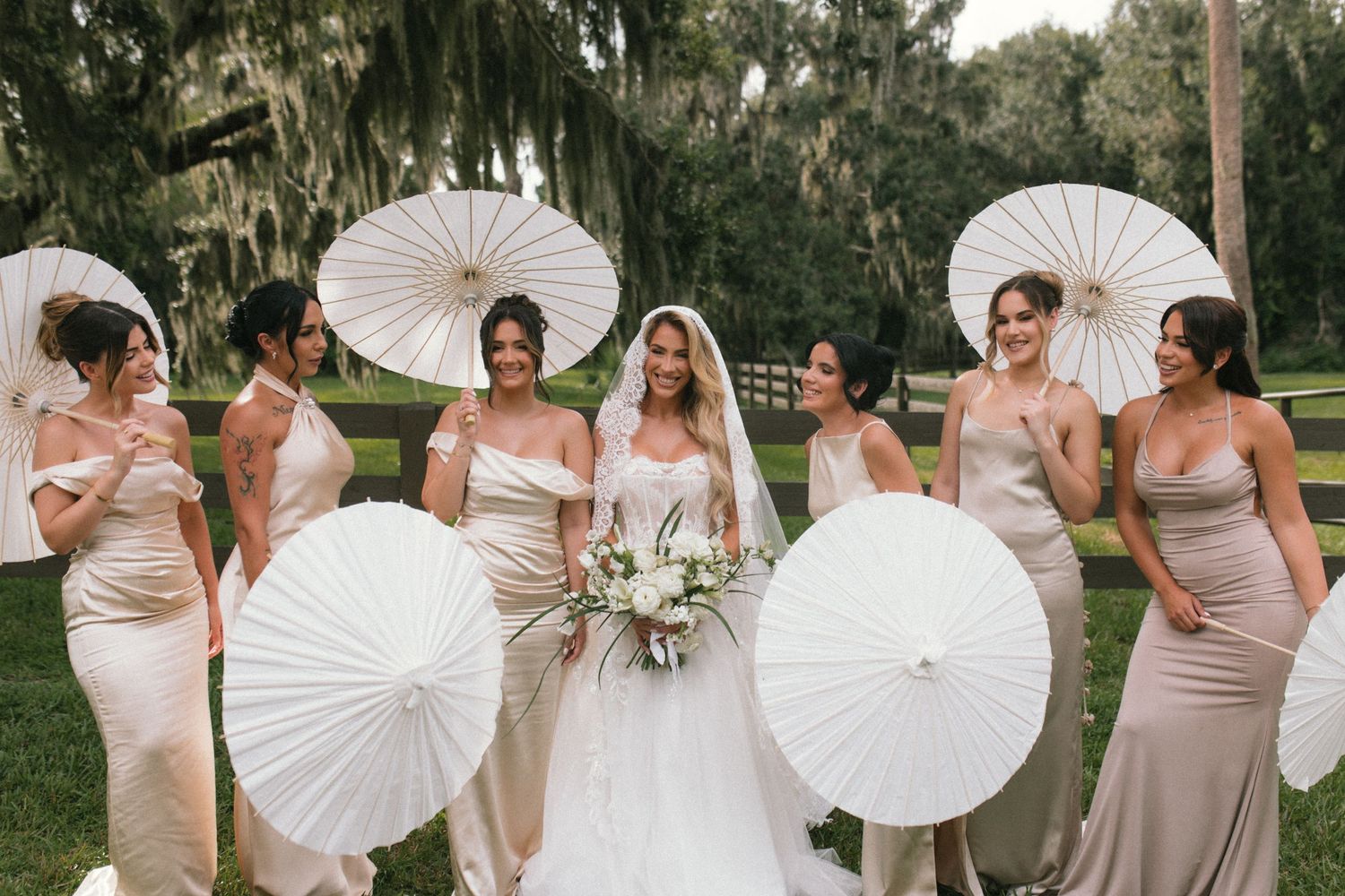 Bridesmaids in blush pink dresses holding white paper parasols pose with bride on grassy lawn under oak trees.