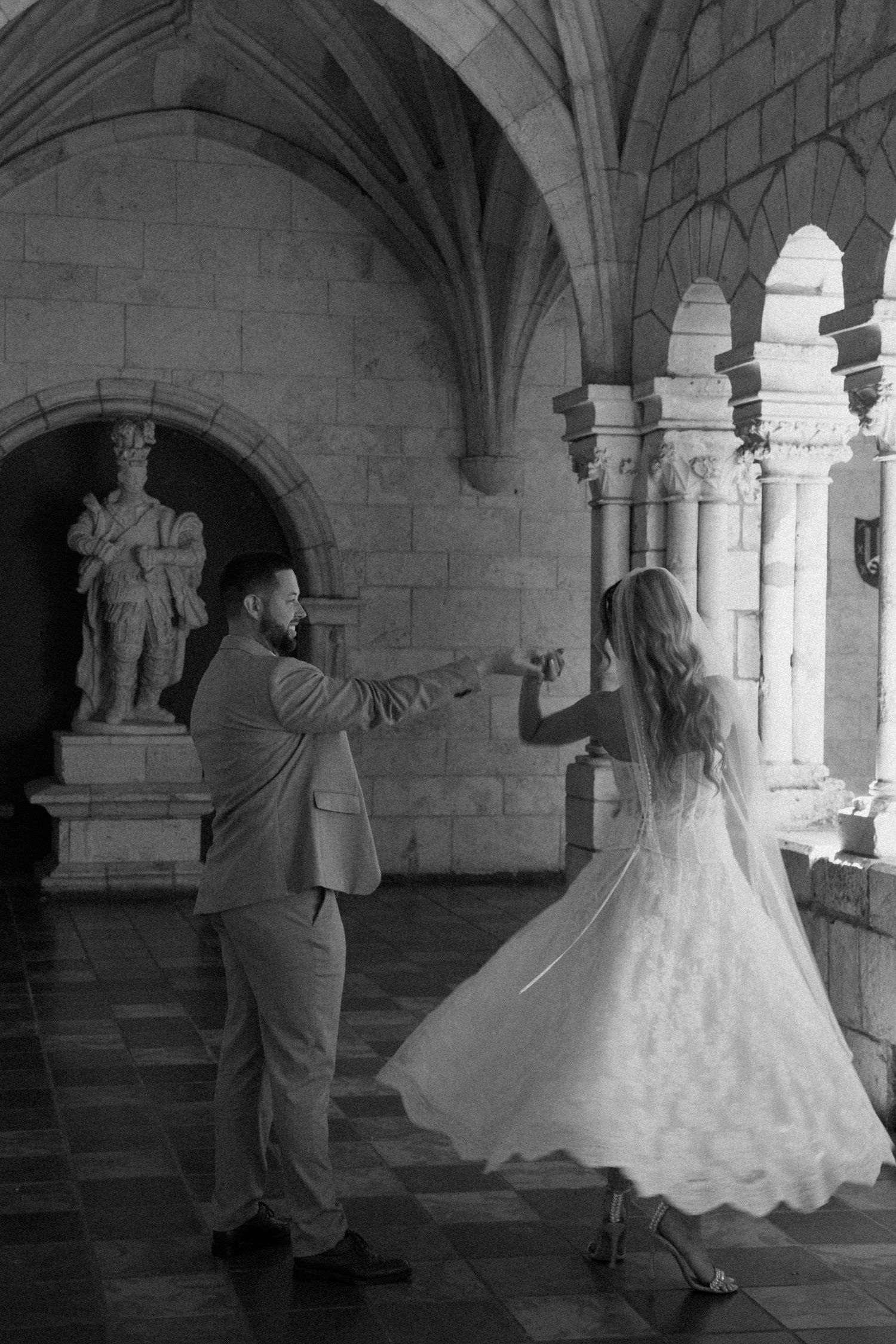 A couple dances together in an elegant stone hallway with vaulted ceilings and arched windows.