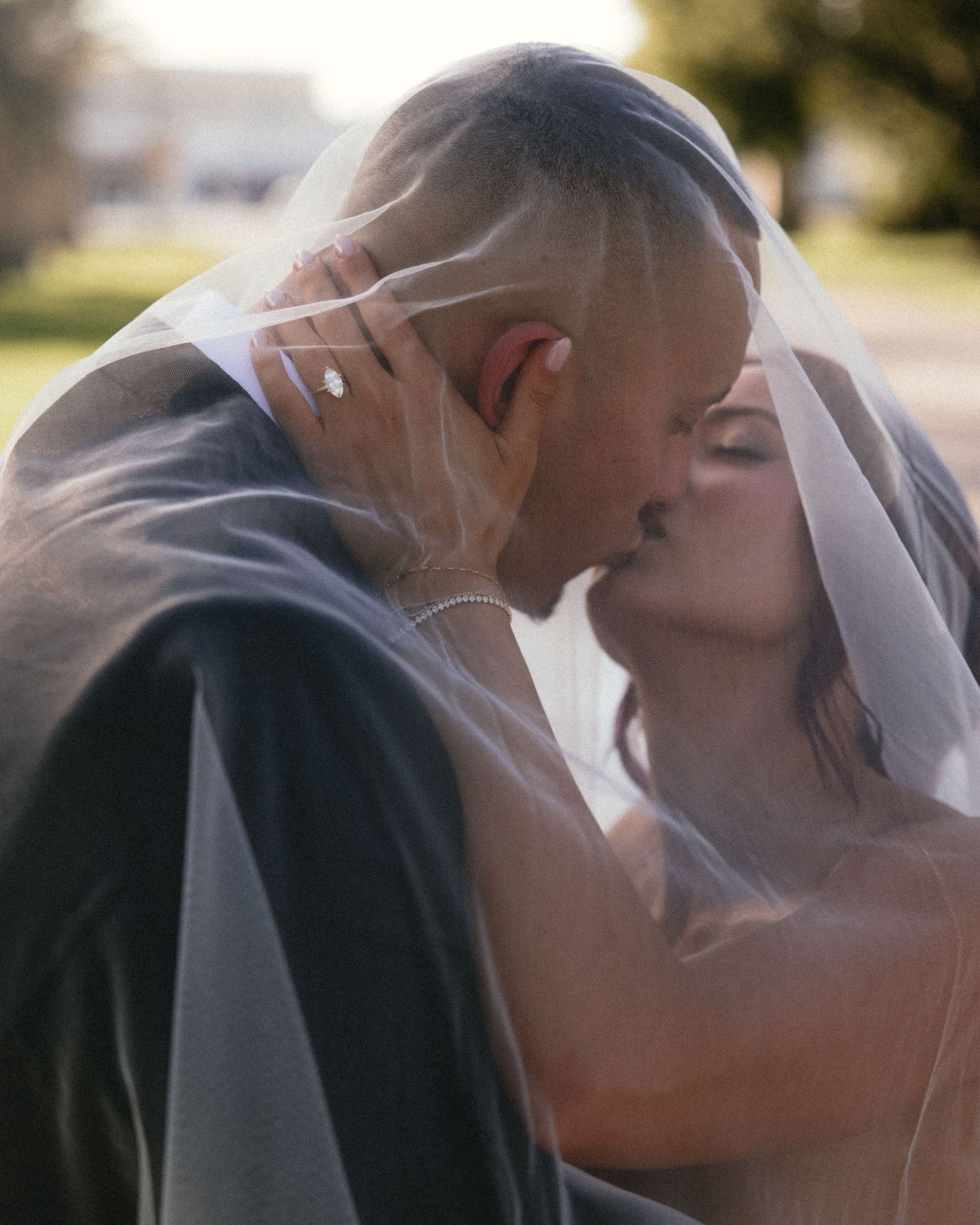 A romantic wedding moment captured as newlyweds share a tender kiss under a flowing white veil in soft sunlight.