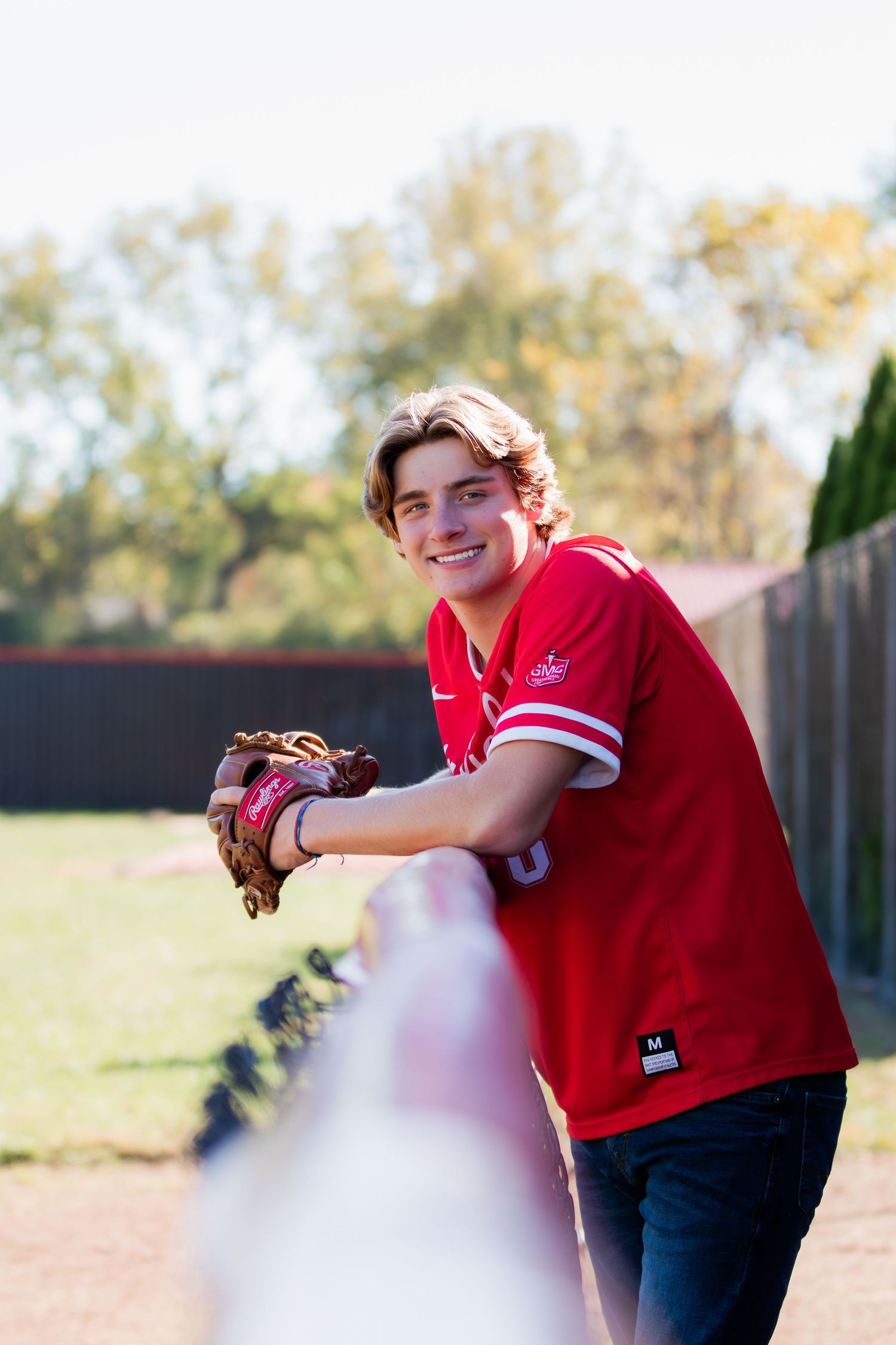 Baseball senior with glove leaning on fence smiling at camera.