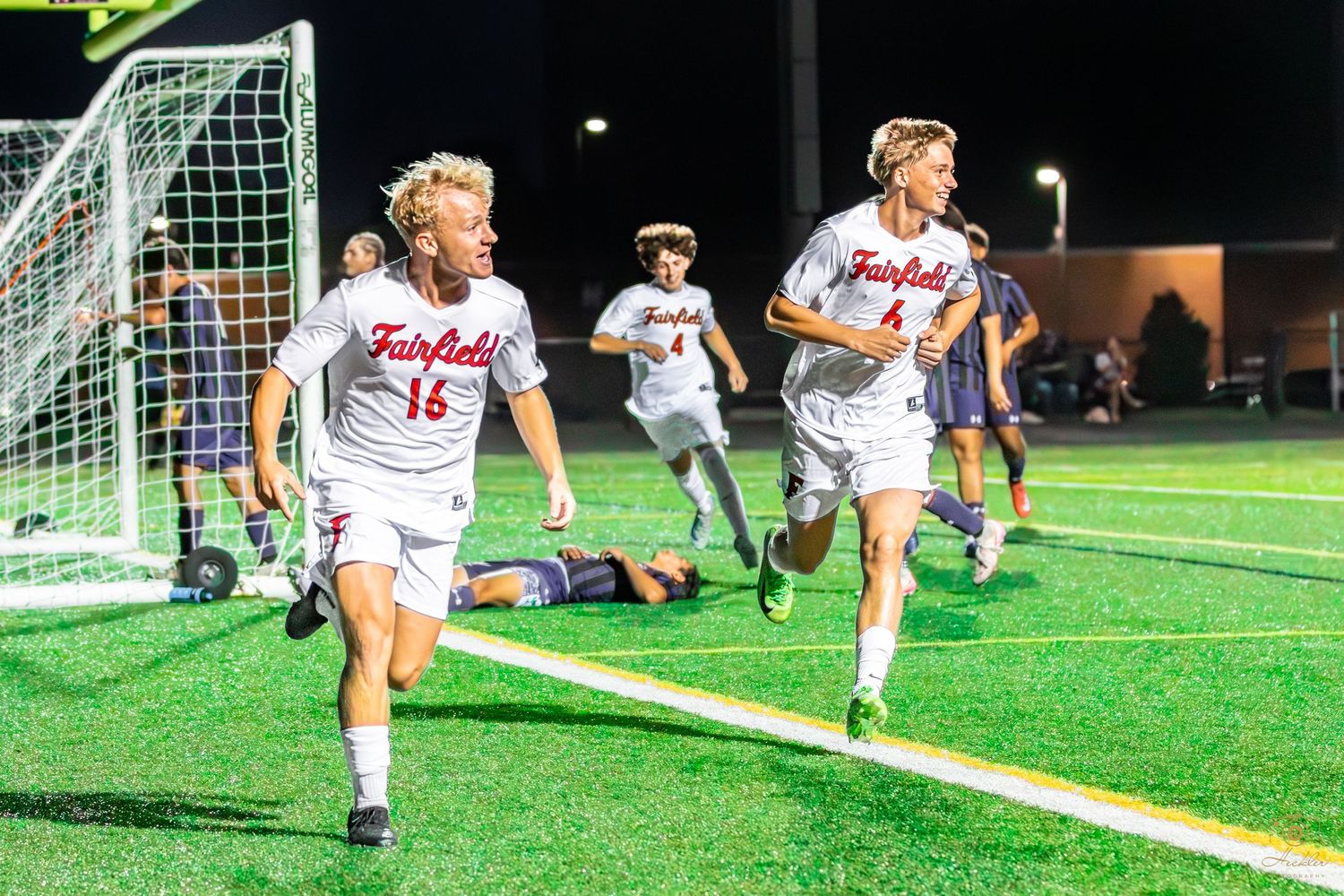 Soccer players celebrating with team after a goal while opposing team is laying on the ground defeated.