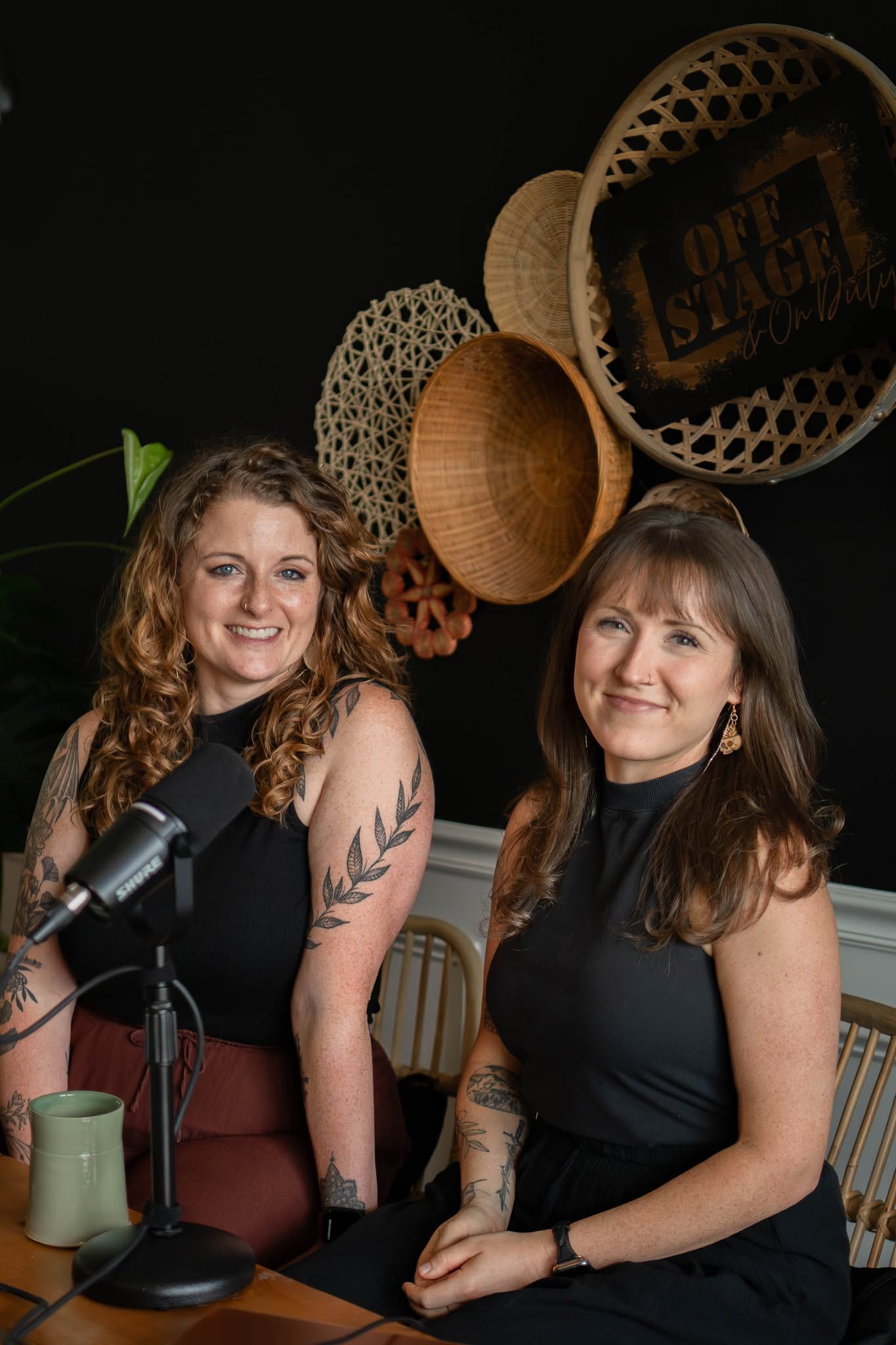 Two women in black tops smile at a podcast microphone, seated before decorative woven baskets and an Off Stage sign.