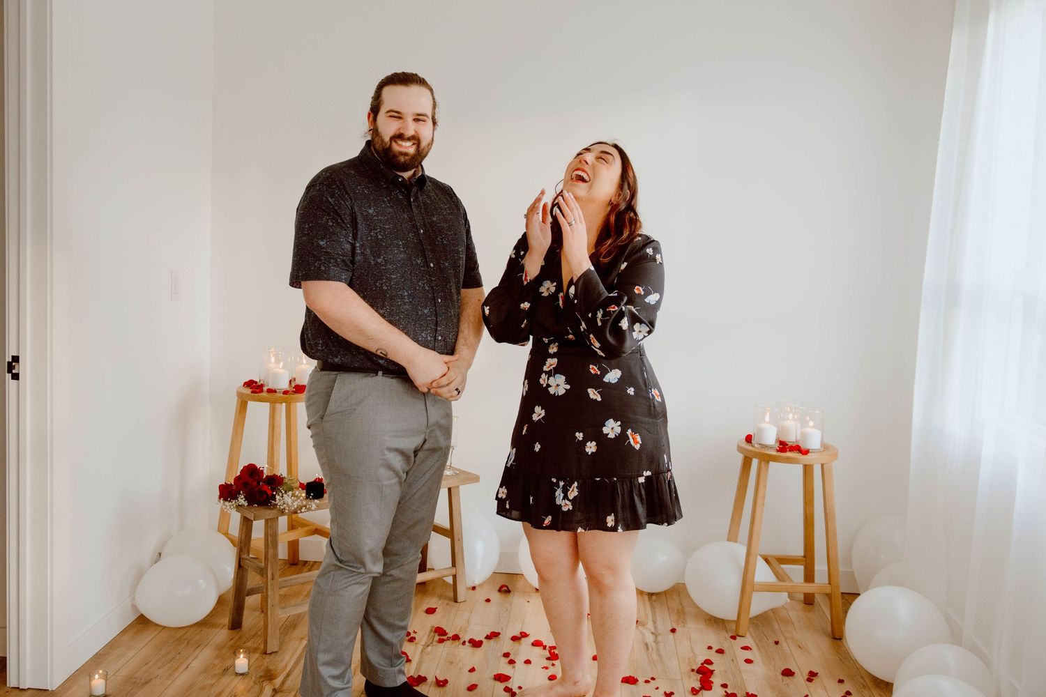 Couple sharing joyful moment surrounded by balloons and rose petals in bright indoor setting.