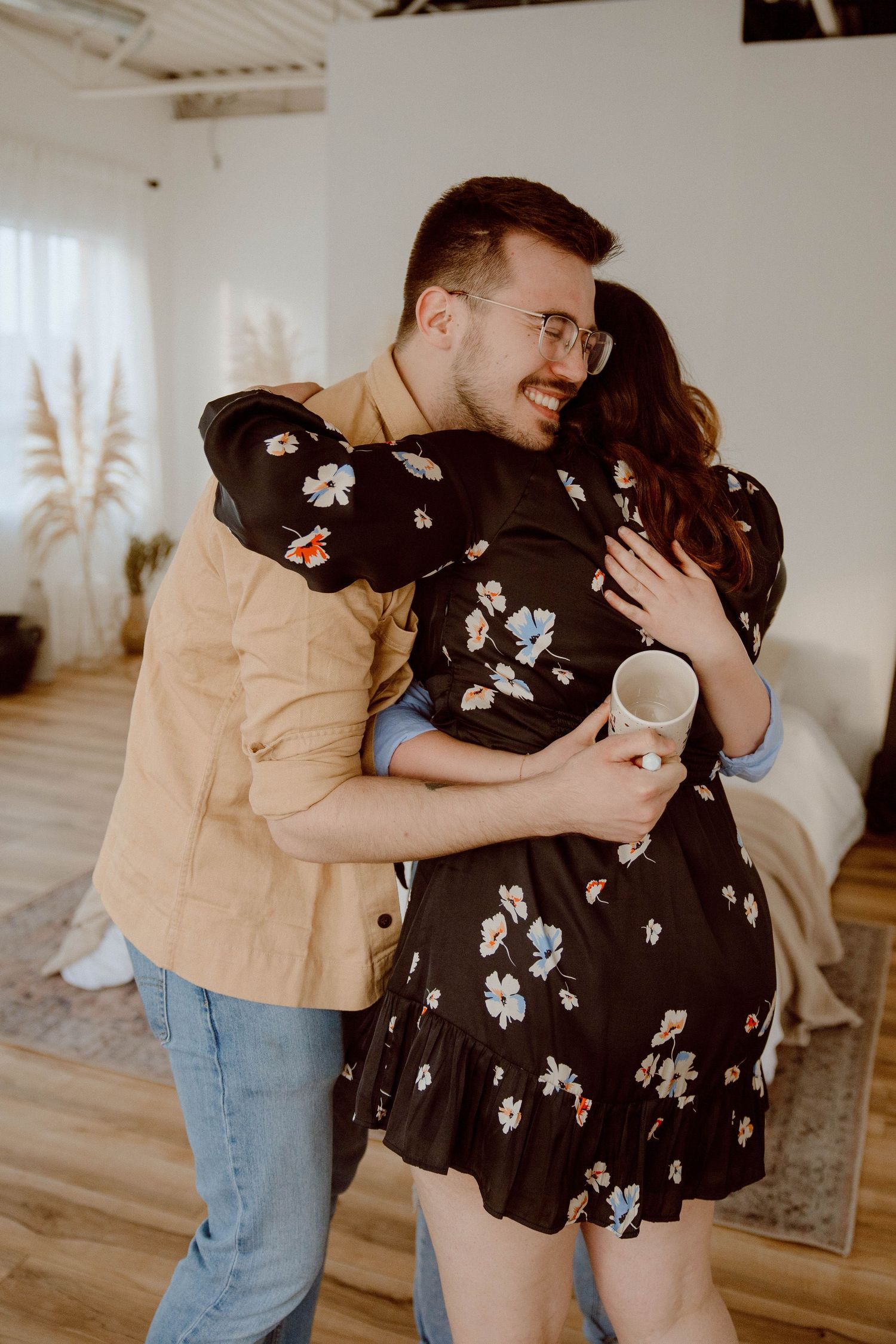 Couple embracing in casual attire against white wall with decorative pampas grass.