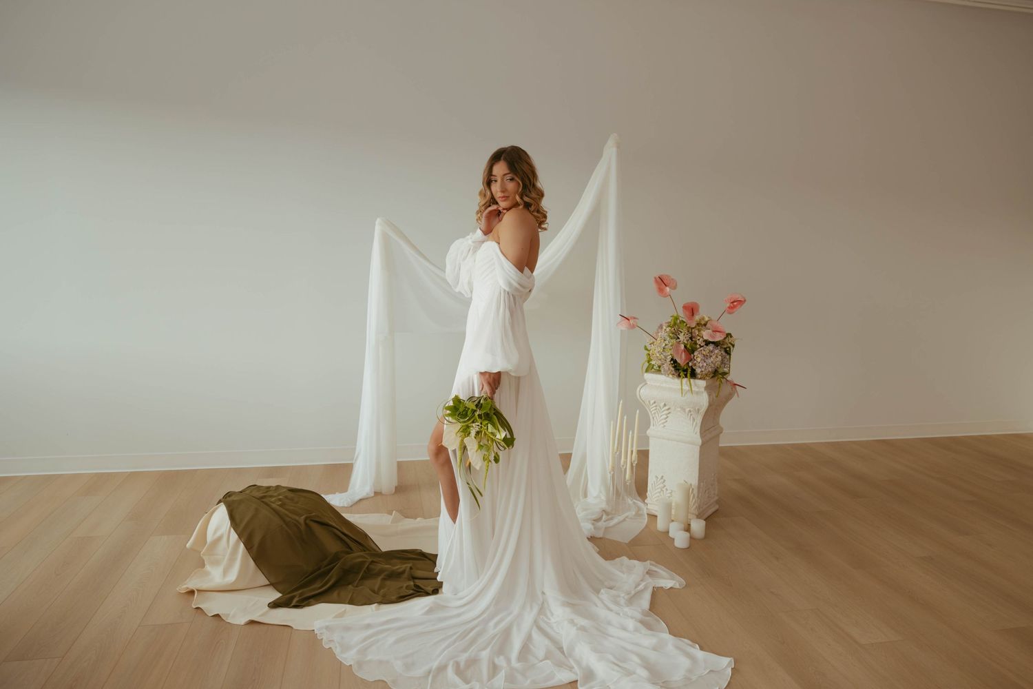 Bride posing in elegant white wedding gown with flowing train against minimalist backdrop.