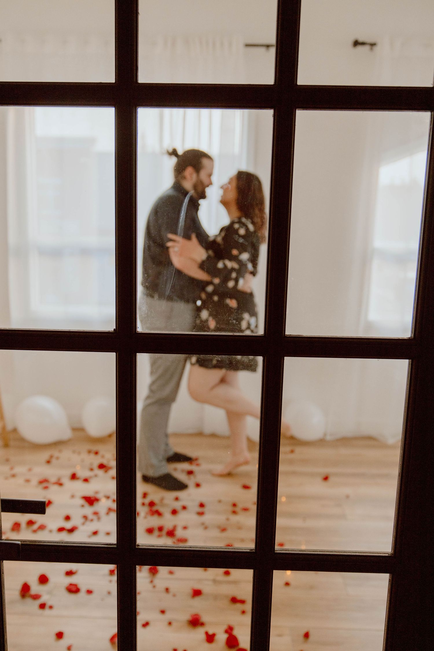 View through window panes of couple in room with scattered rose petals and balloons.