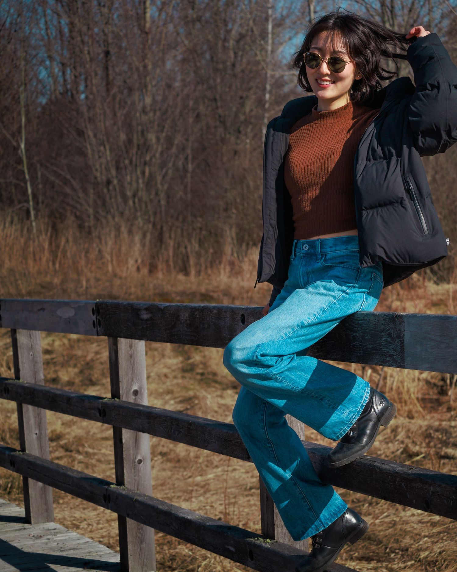 Casual winter fashion photo of someone in puffer jacket and blue jeans sitting on wooden fence.