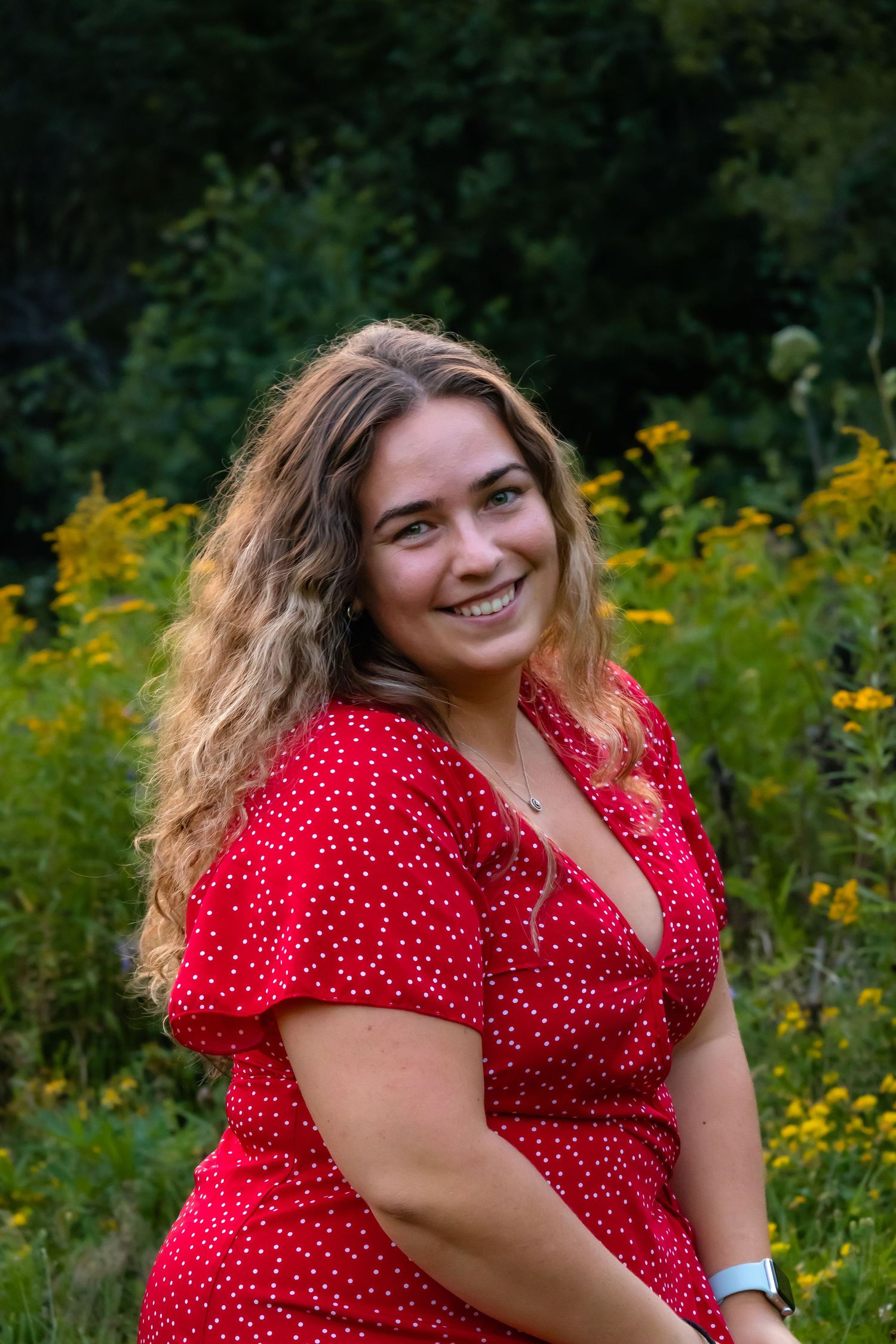 Smiling woman in red polka dot dress posing outdoors with yellow wildflowers in background.