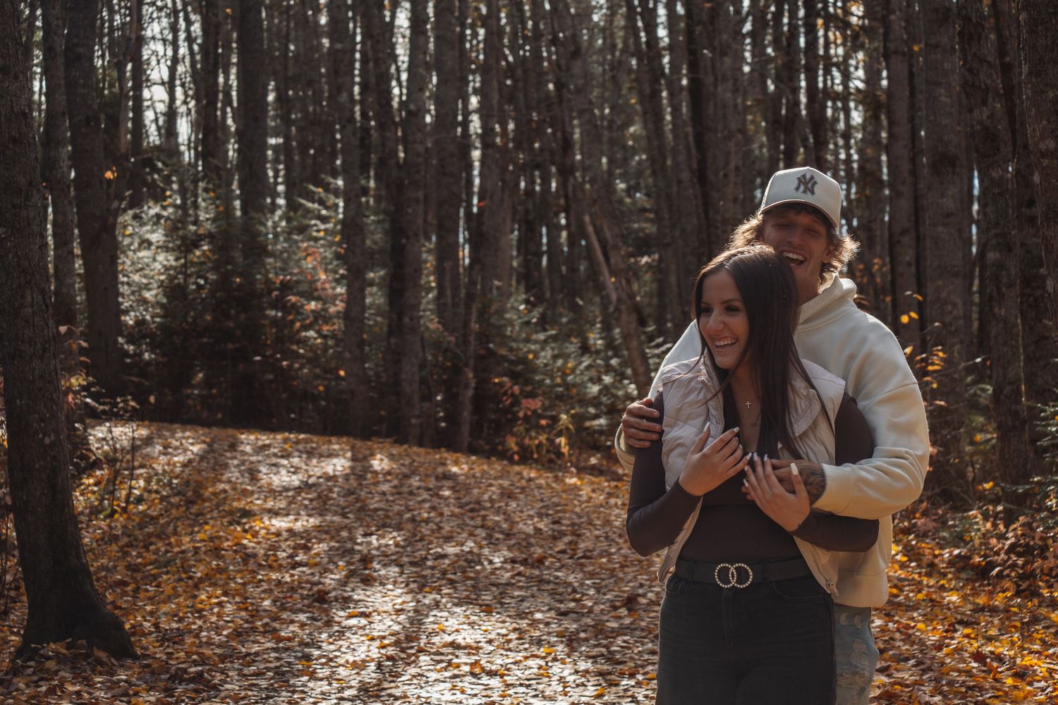 A couple embraces in a romantic autumn forest setting with sunlight filtering through the trees.