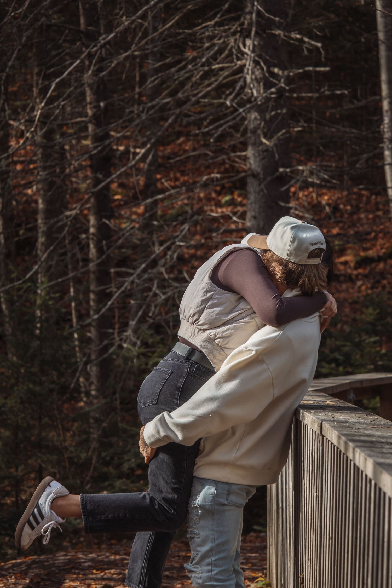 Two people share a romantic embrace while leaning against a wooden railing in an autumn forest setting.