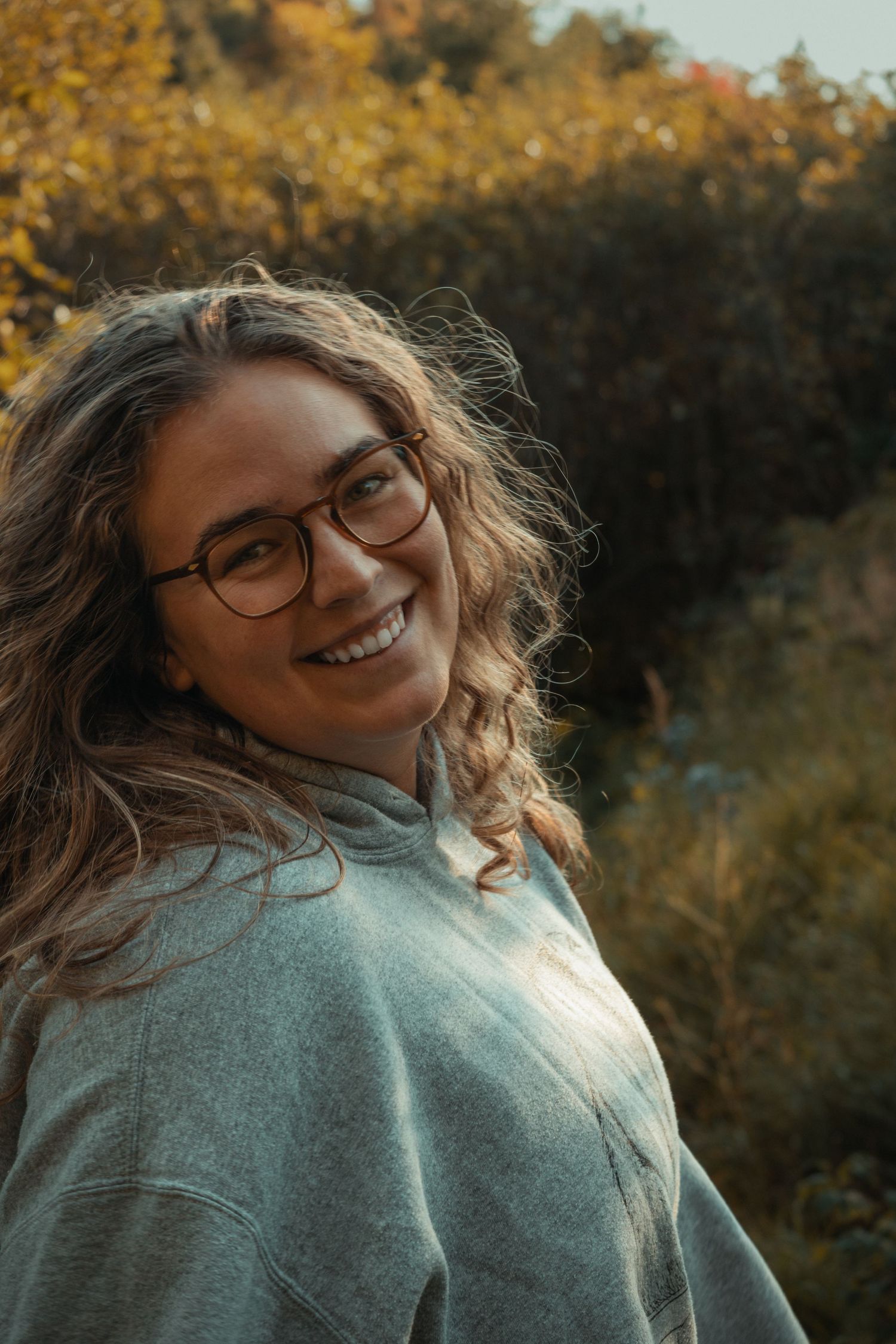 A person in a gray sweater laughs joyfully while posing outdoors with autumn foliage in the background.