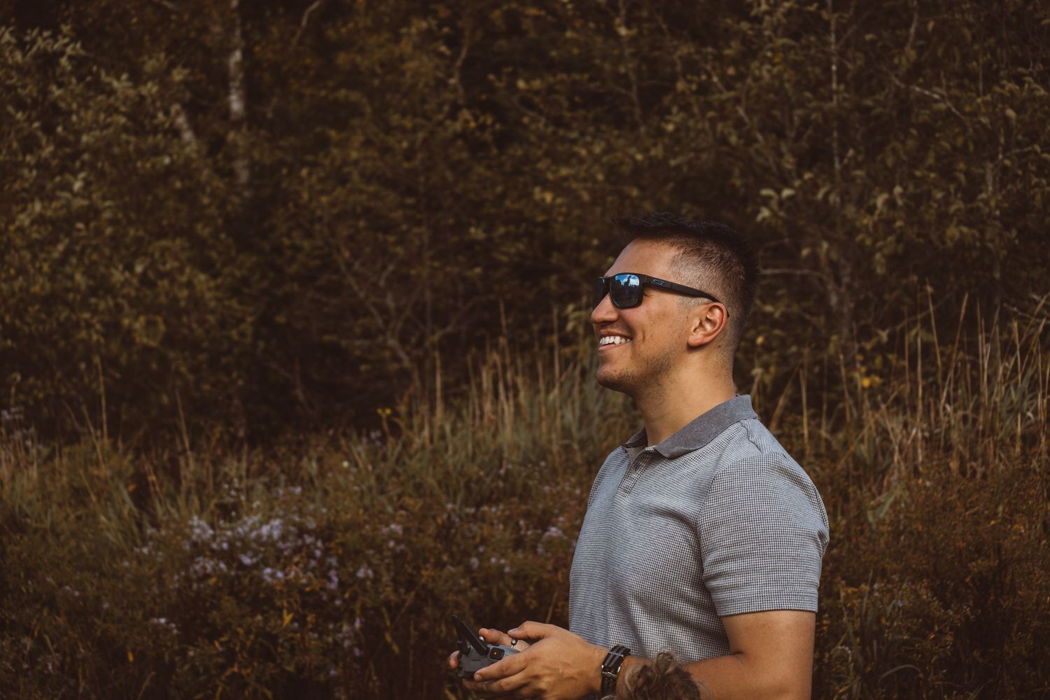 A person in a gray t-shirt and sunglasses stands in a wooded area holding a phone and looking to the side.