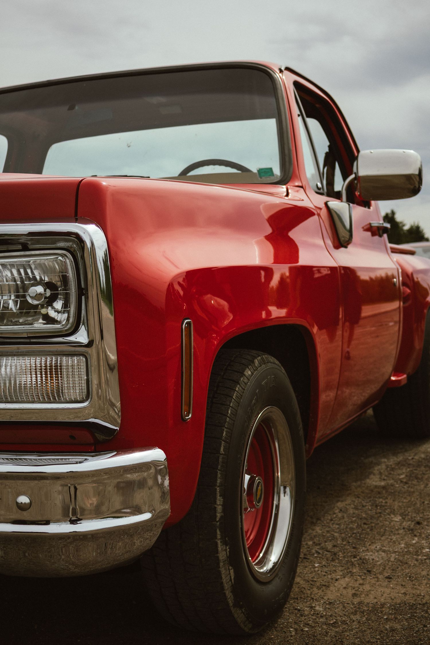 A shiny red classic pickup truck with chrome accents and red rims parked outdoors under a cloudy sky.