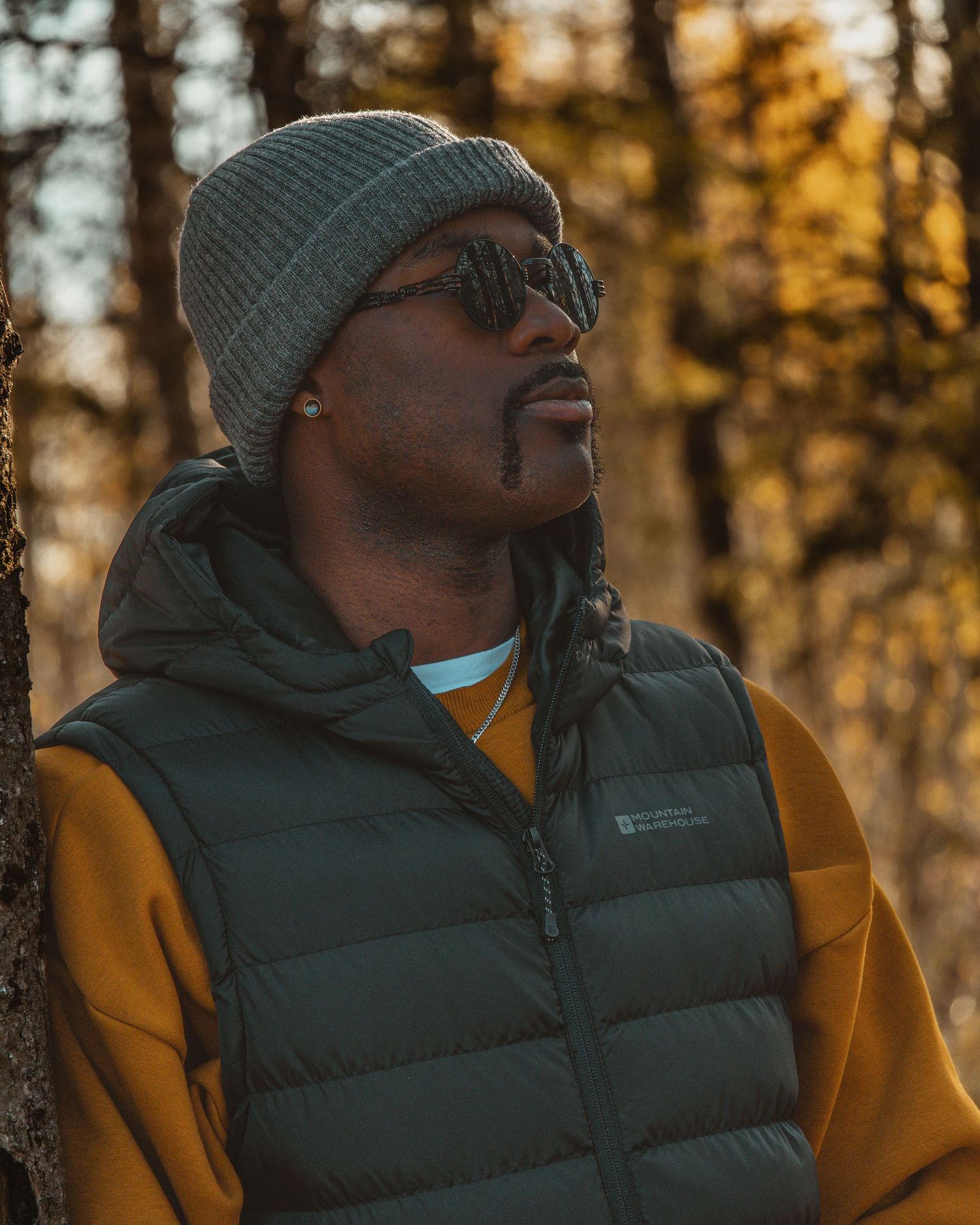 Person wearing sunglasses and beanie looks upward thoughtfully in autumn forest during golden hour sunset.