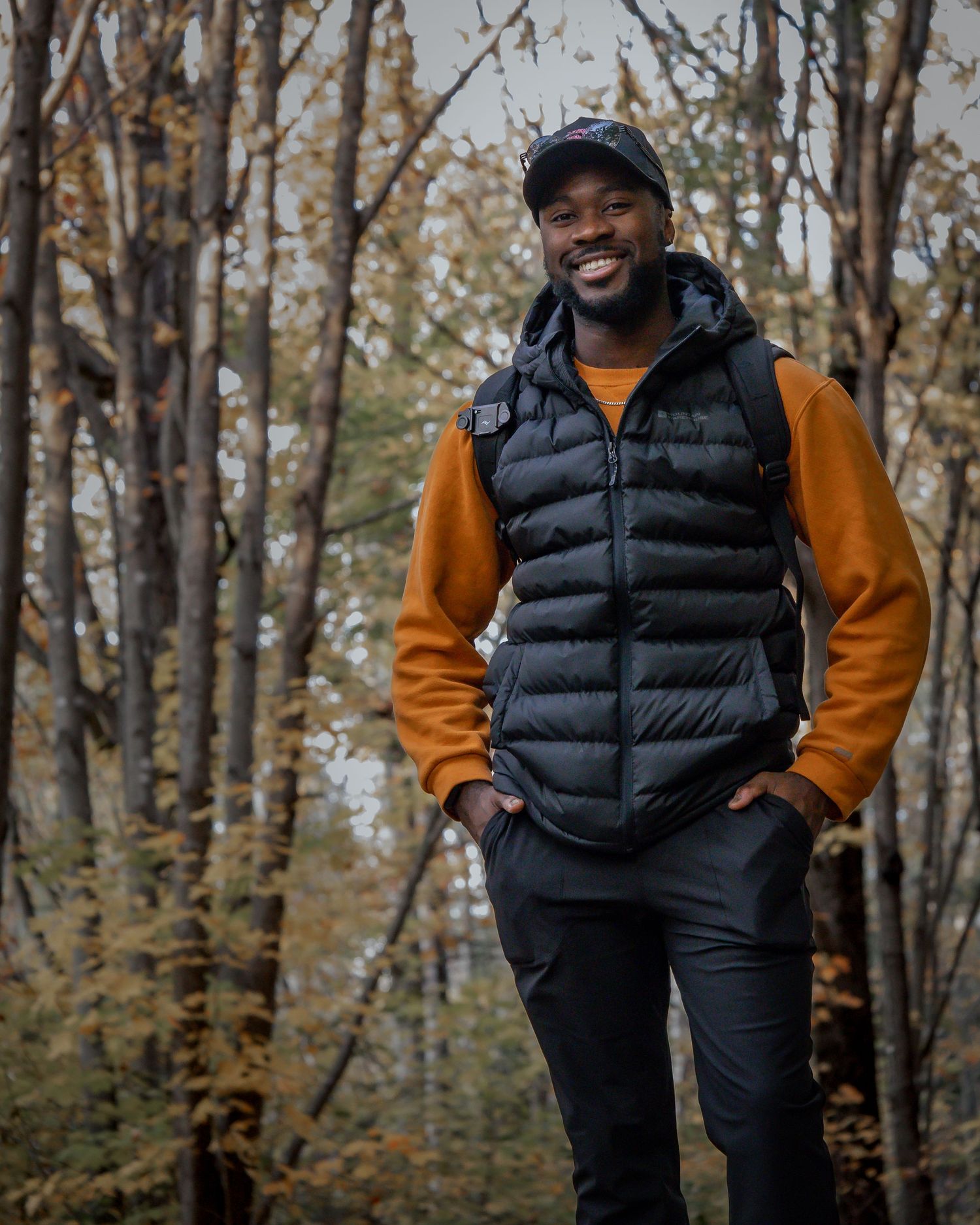 Person wearing orange sweater and black puffer vest standing among bare autumn trees in forest.
