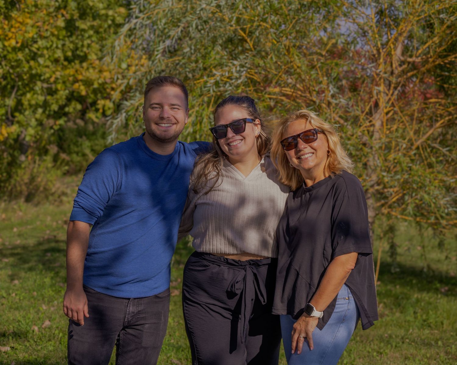 Family group poses together outdoors in casual attire against autumn foliage background.