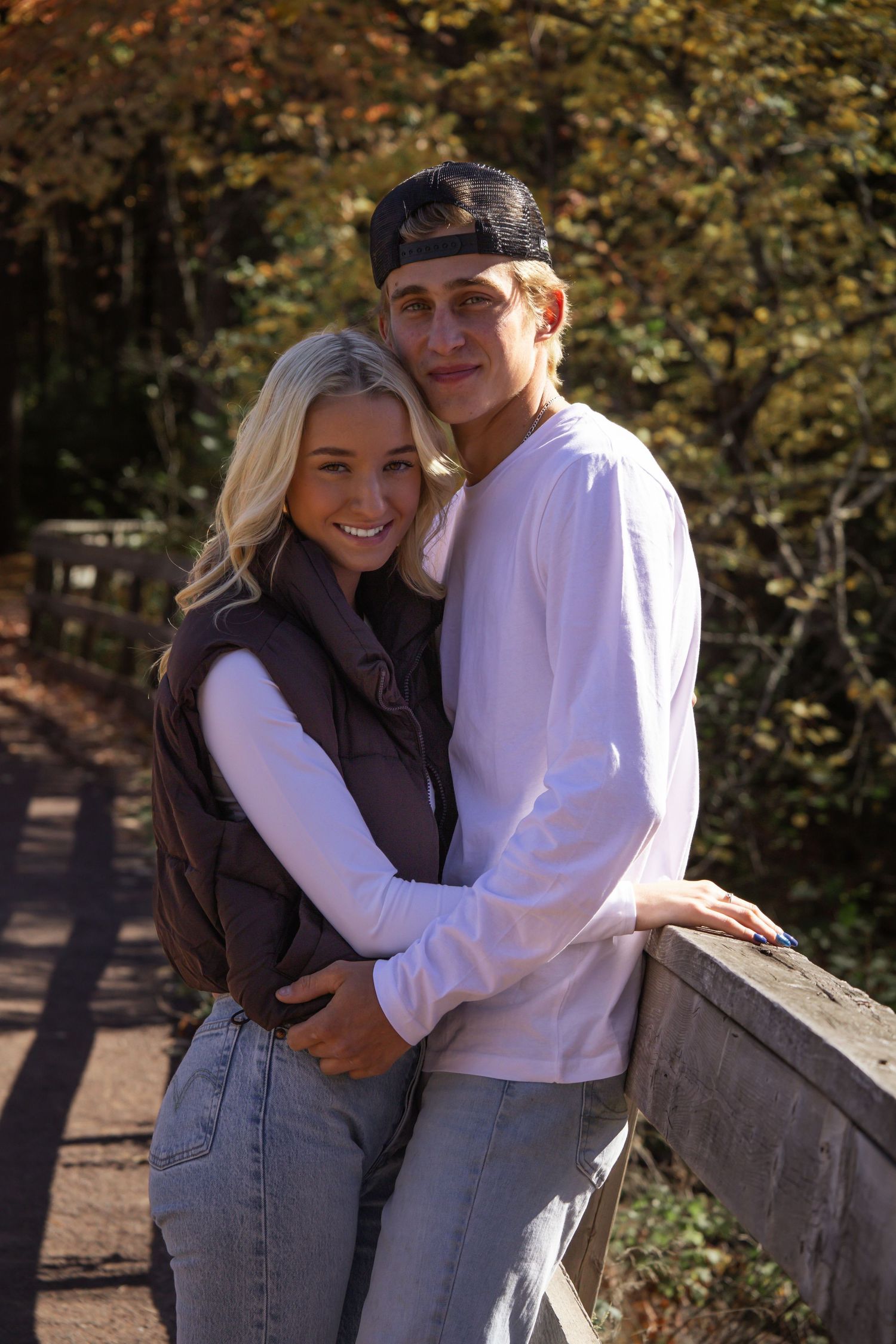 A couple embraces and smiles together on a bridge in an autumn park setting with trees in the background.