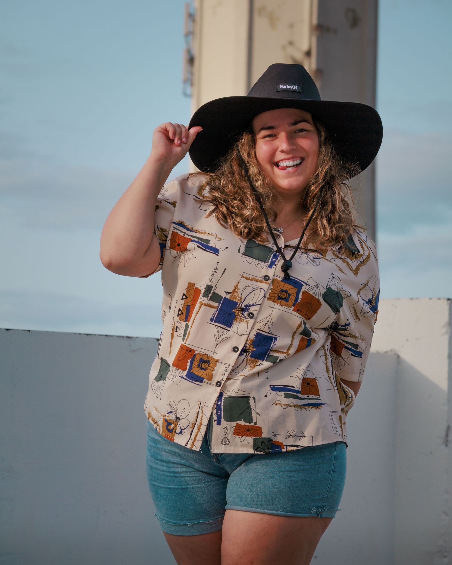 Person in black hat and patterned shirt posing against white wall outdoors.