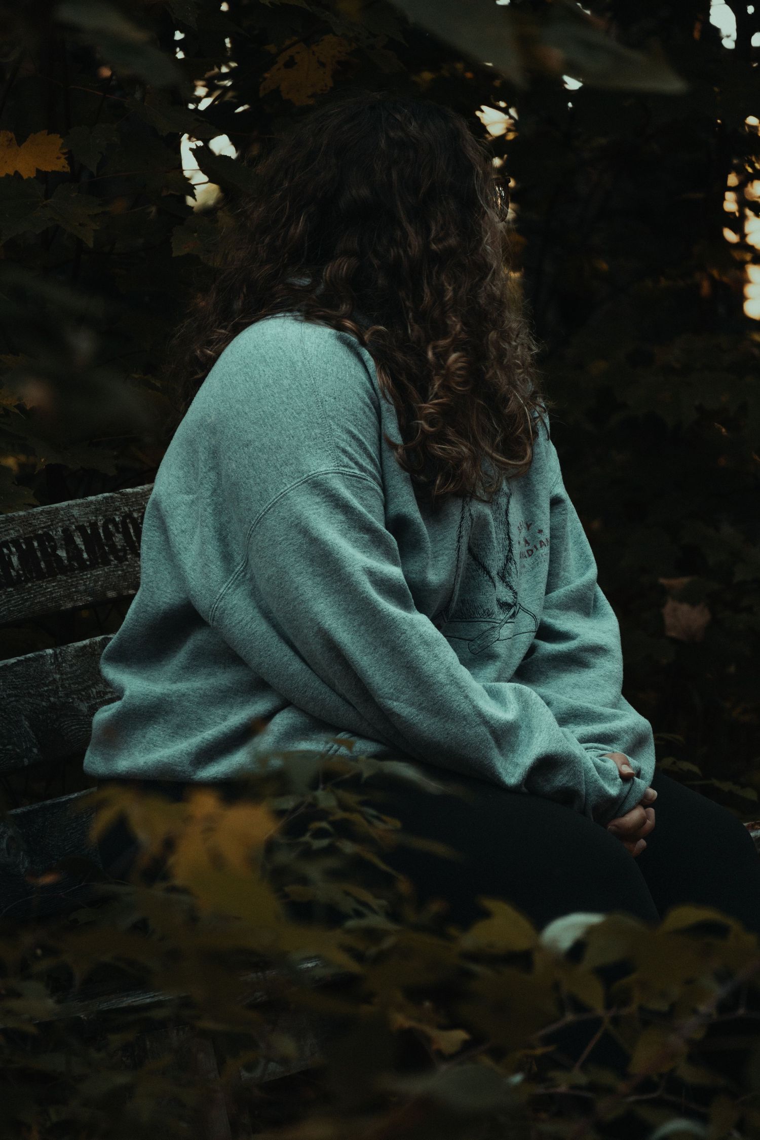 A person wearing a teal sweatshirt sits in a dark moody setting with autumn leaves in the background.