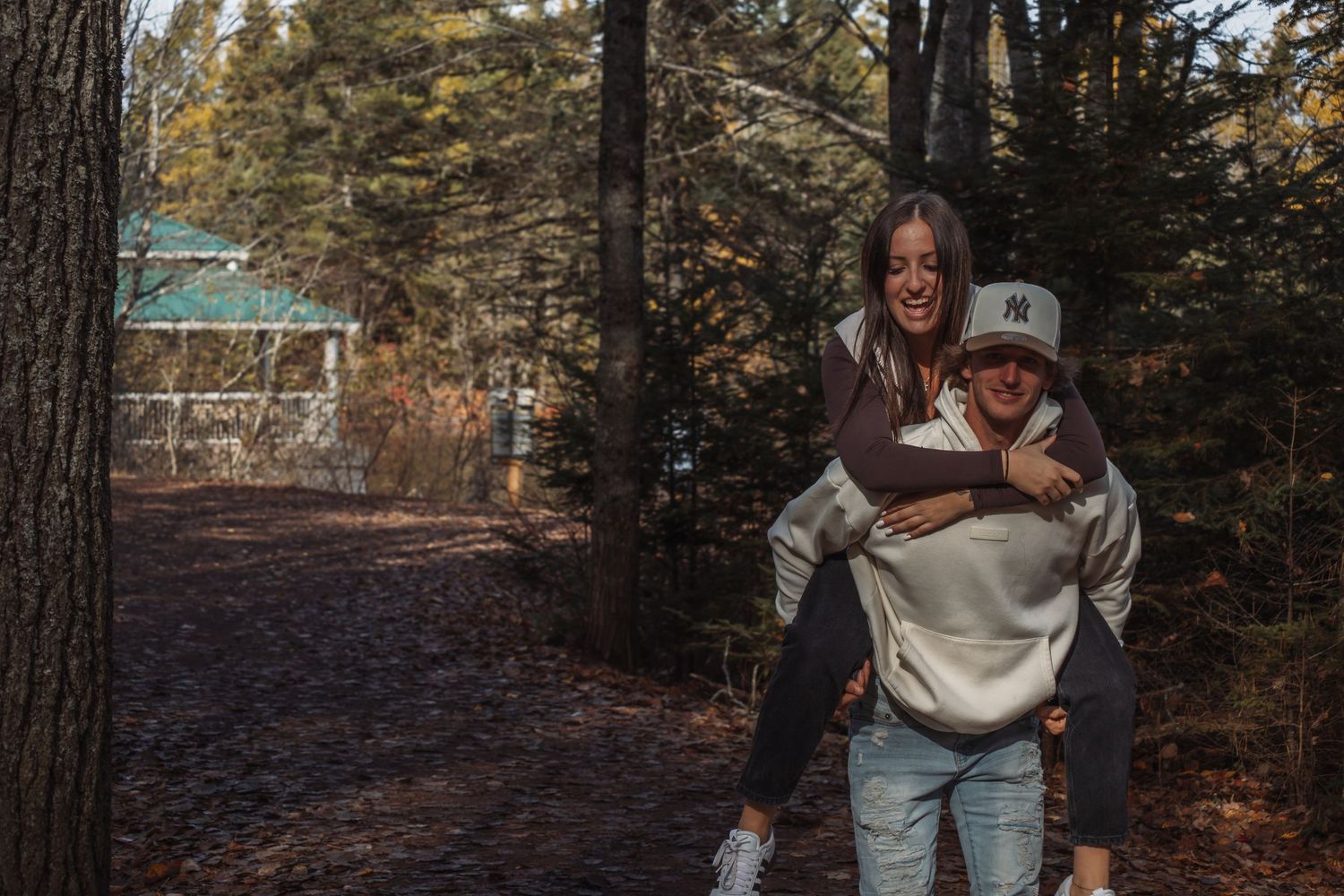 A playful piggyback ride moment captured between two people on a forest trail surrounded by tall pine trees.